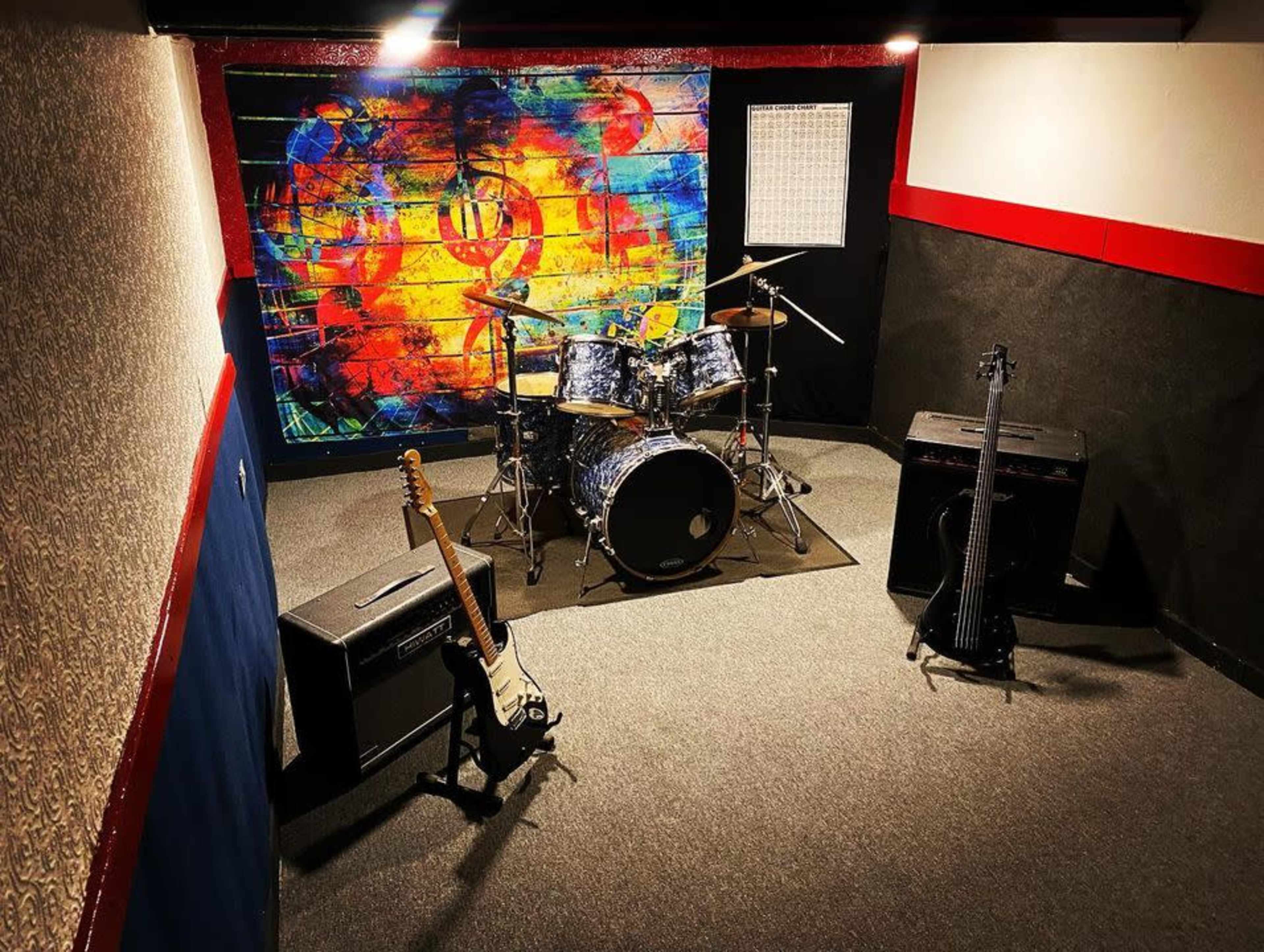 The image shows a music rehearsal studio featuring a drum set, an electric guitar, an amplifier, and a colorful mural on the back wall.