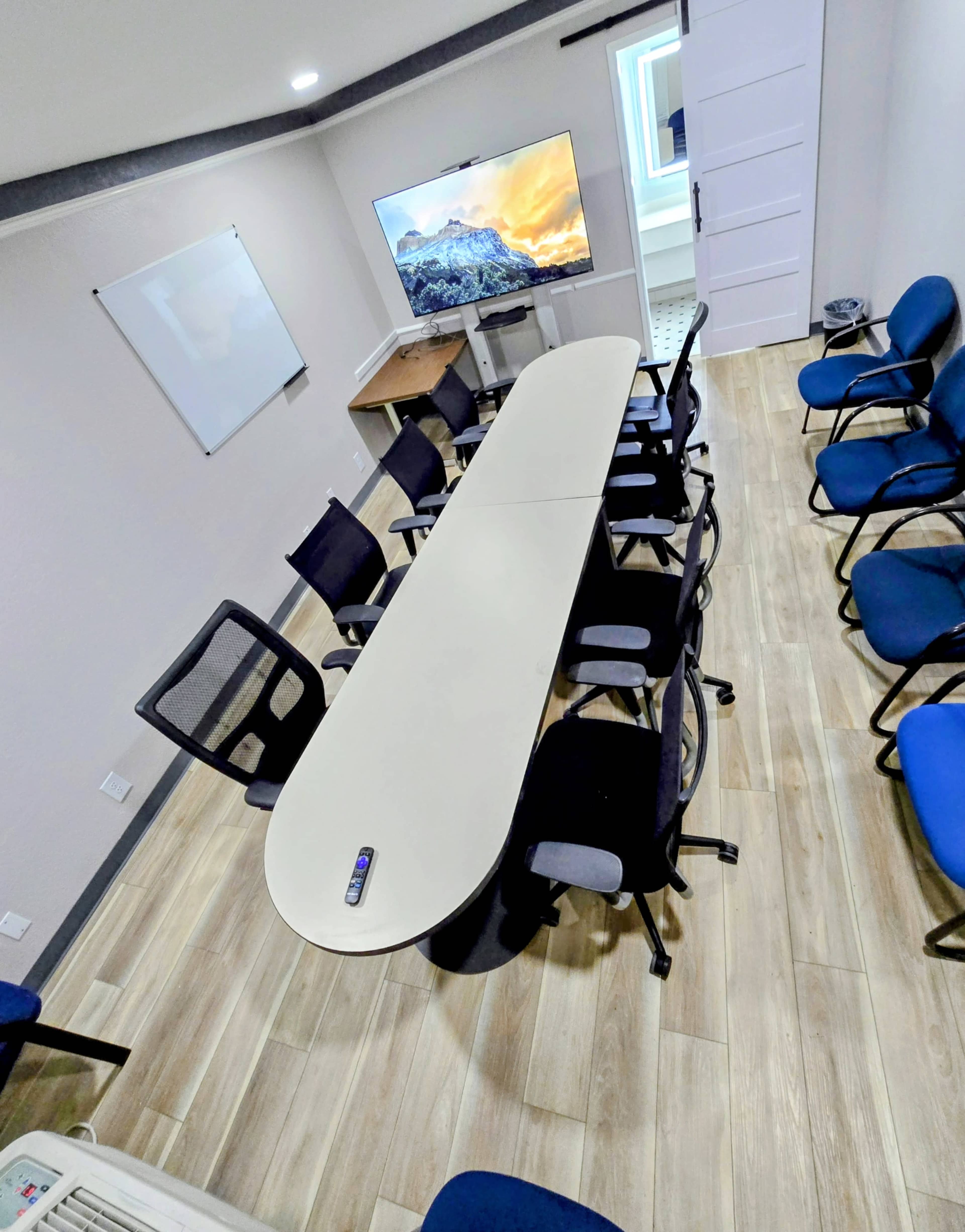 A long, oval conference table surrounded by several chairs is set up in a modern meeting room with a wall-mounted television and a whiteboard.