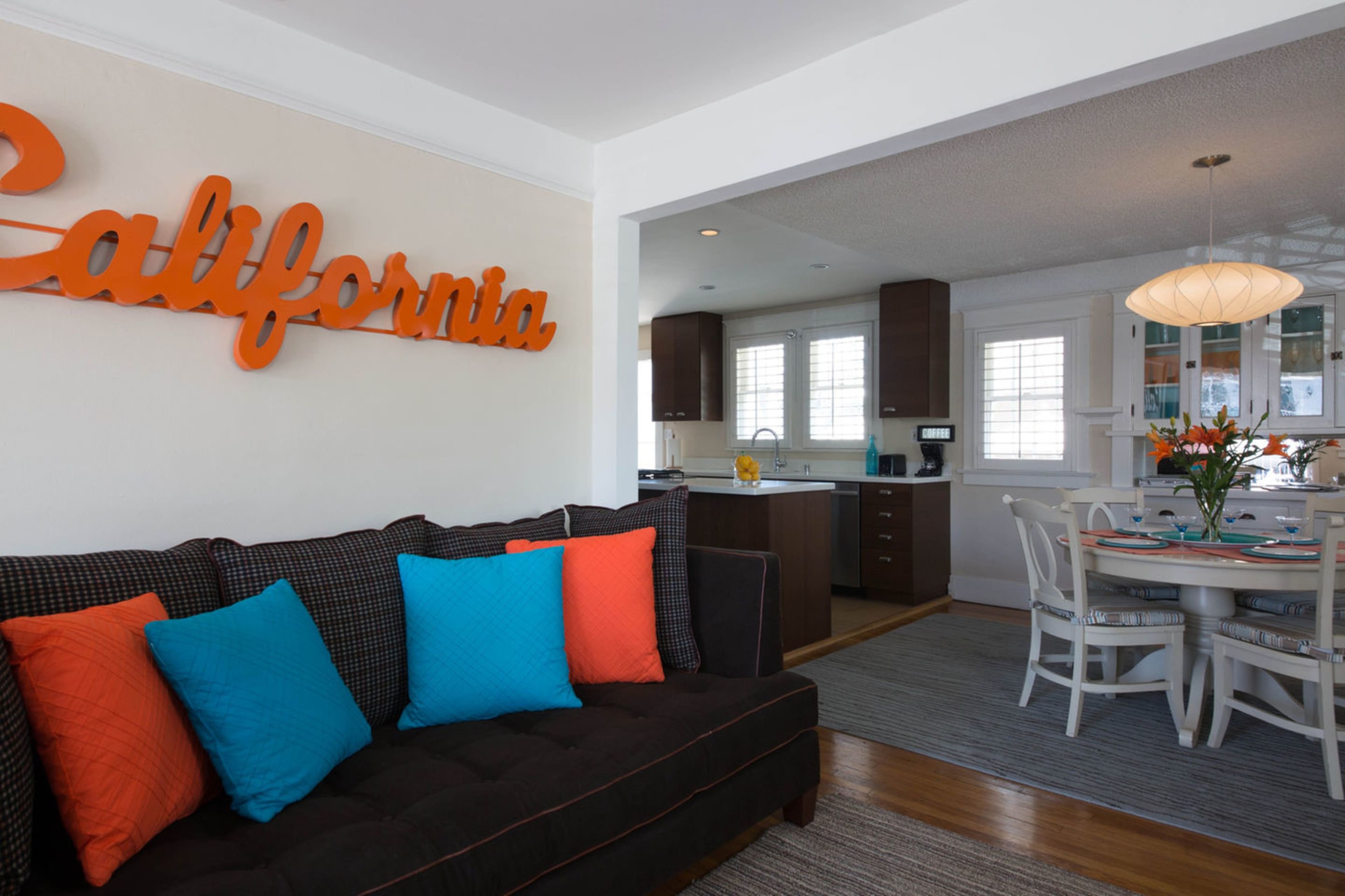 A living room features a dark couch with colorful pillows, a large orange "California" sign on the wall, and an adjacent dining area with a round table and white chairs.