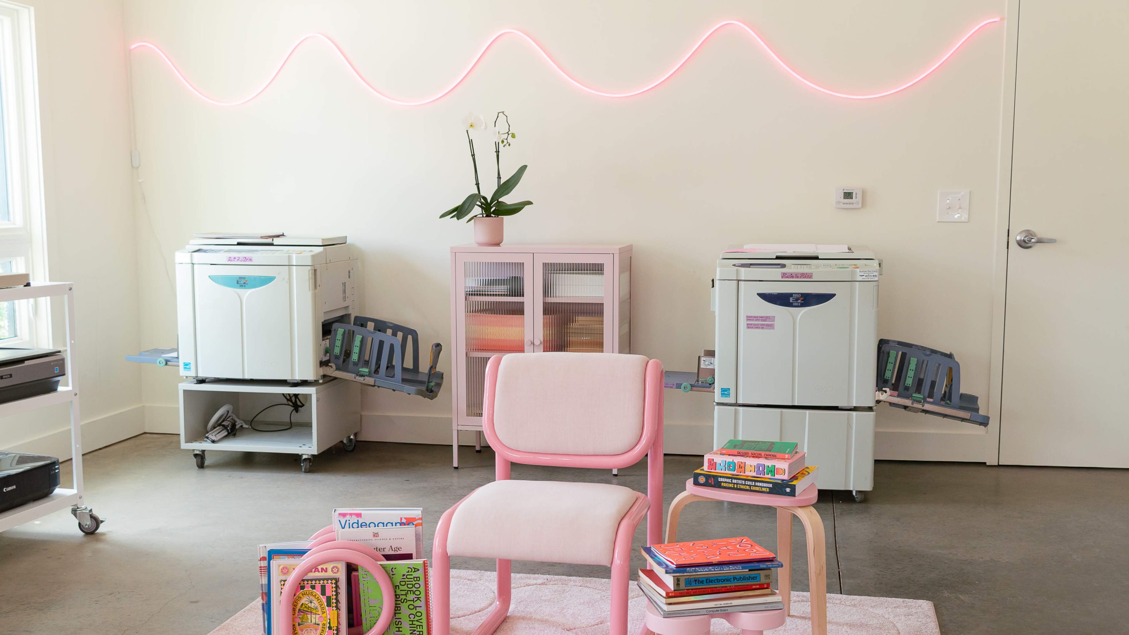 The image shows two photocopiers on mobile stands against a wall with a pink neon wave light, accompanied by a pink chair and a stack of colorful books on a rug.