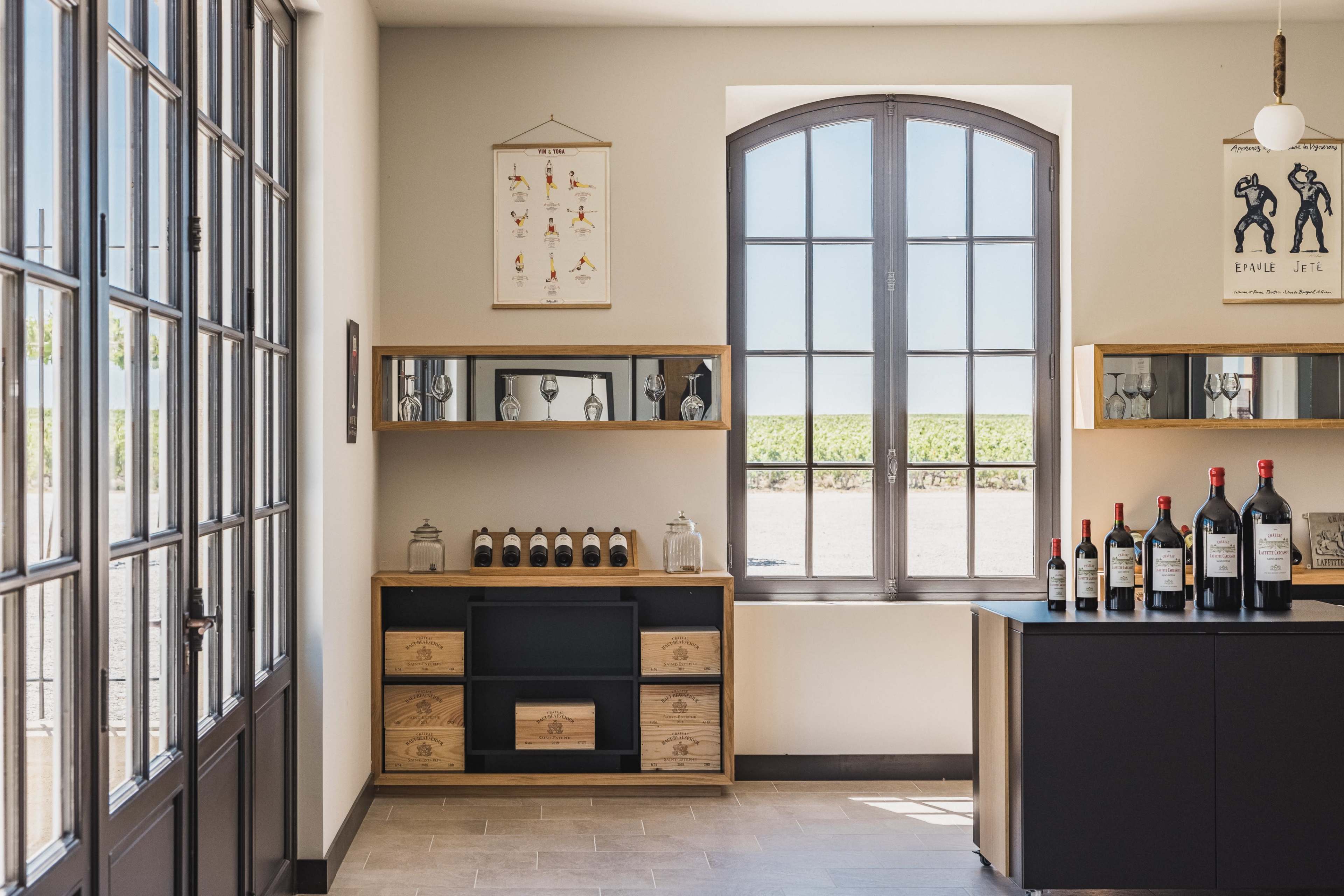 The image shows a modern interior of a winery tasting room with large windows, displaying bottles of wine on a shelf and a counter.