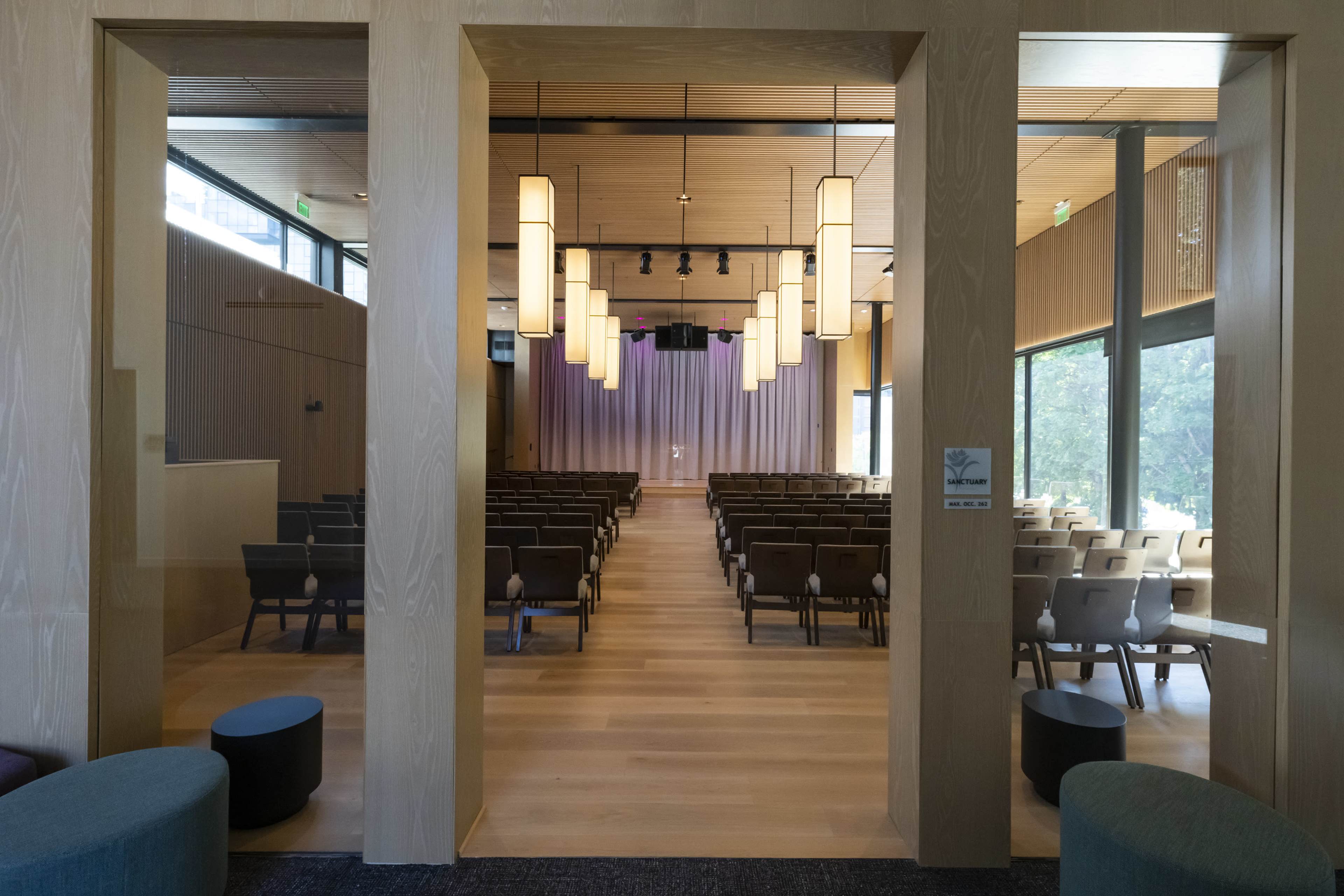 A spacious auditorium with rows of chairs facing a stage, illuminated by pendant lights and framed by wooden walls.