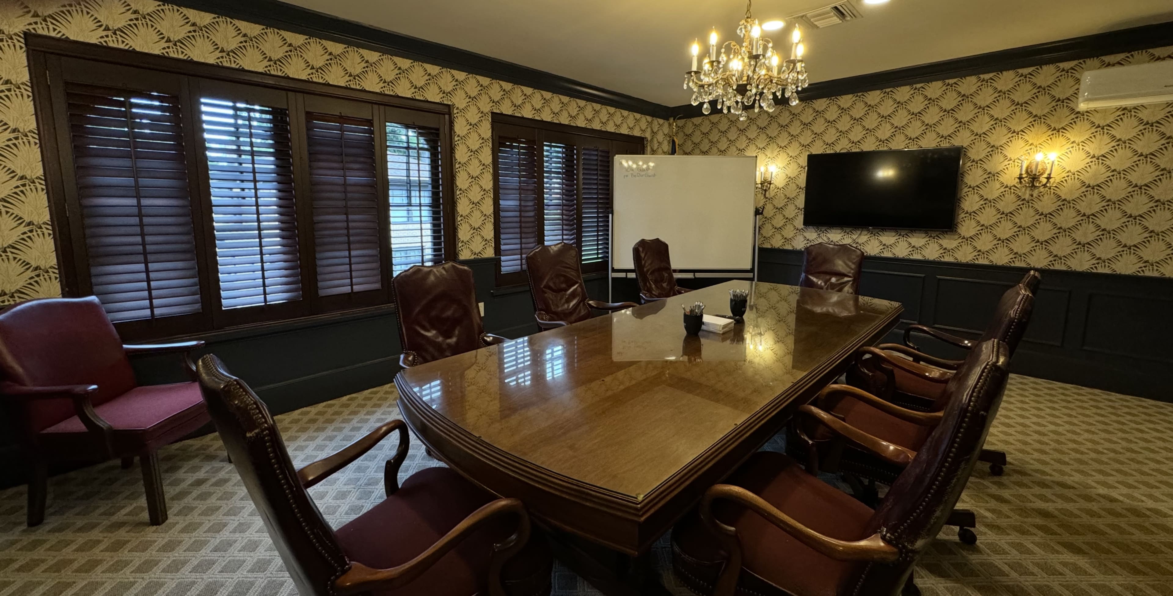 The image shows a conference room featuring a large wooden table surrounded by leather executive chairs, with a whiteboard, a television screen, and ornate chandeliers illuminating the space.