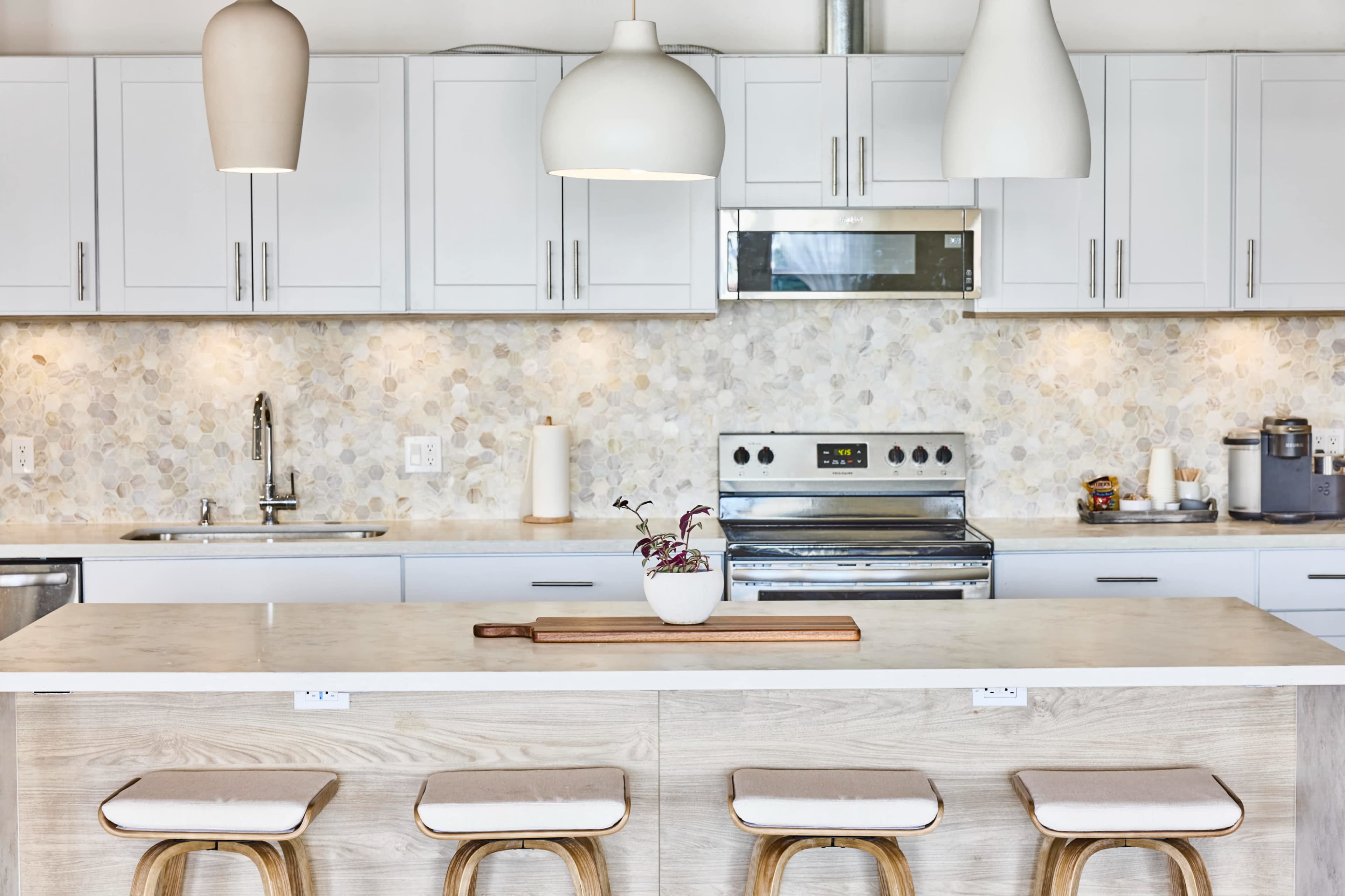The image shows a modern kitchen featuring white cabinetry, a marble-patterned backsplash, a stainless steel stove, and barstools at an island with a wooden cutting board on top.