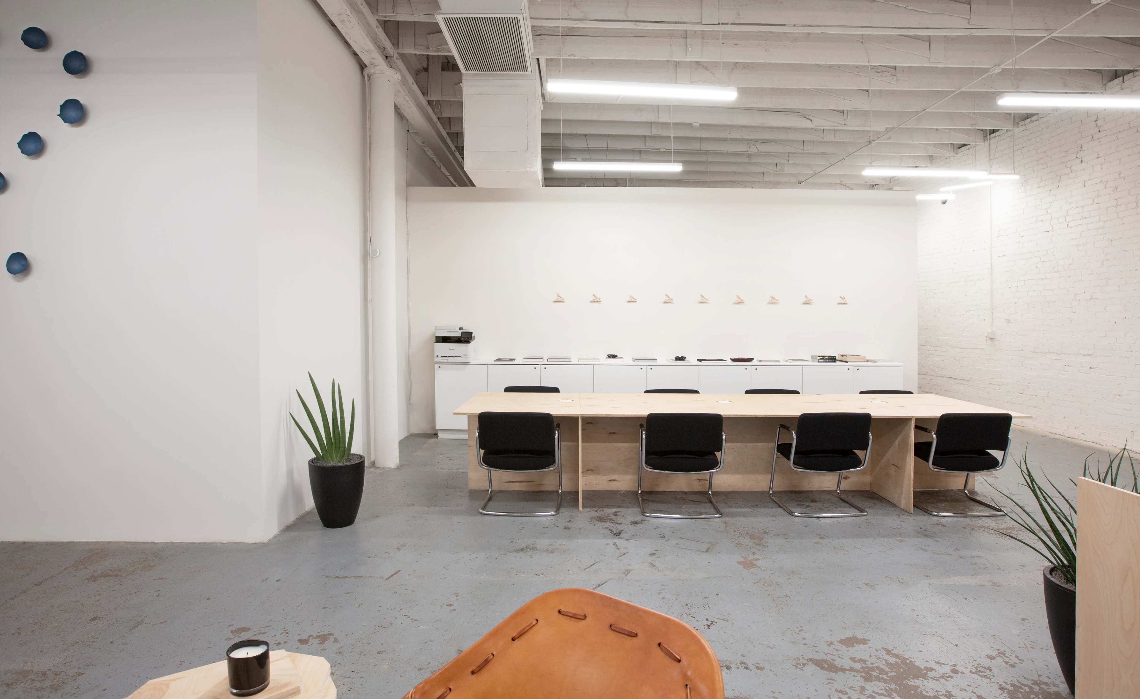 A spacious, minimalist office interior with a long wooden table, black chairs, and a light-colored wall adorned with small lights.