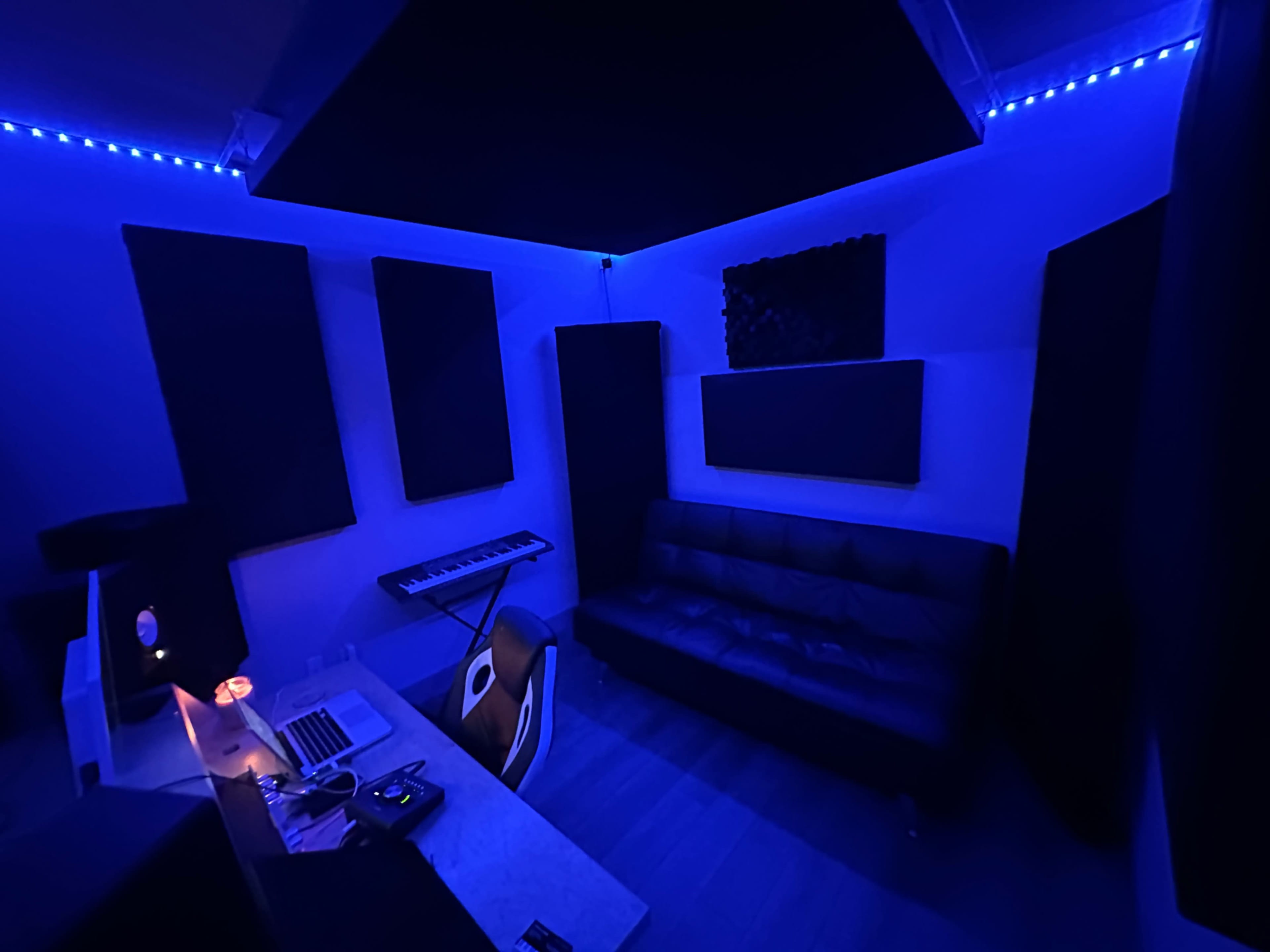 A dimly lit music studio with blue LED lights, featuring a keyboard, a black sofa, and a desk with a laptop.
