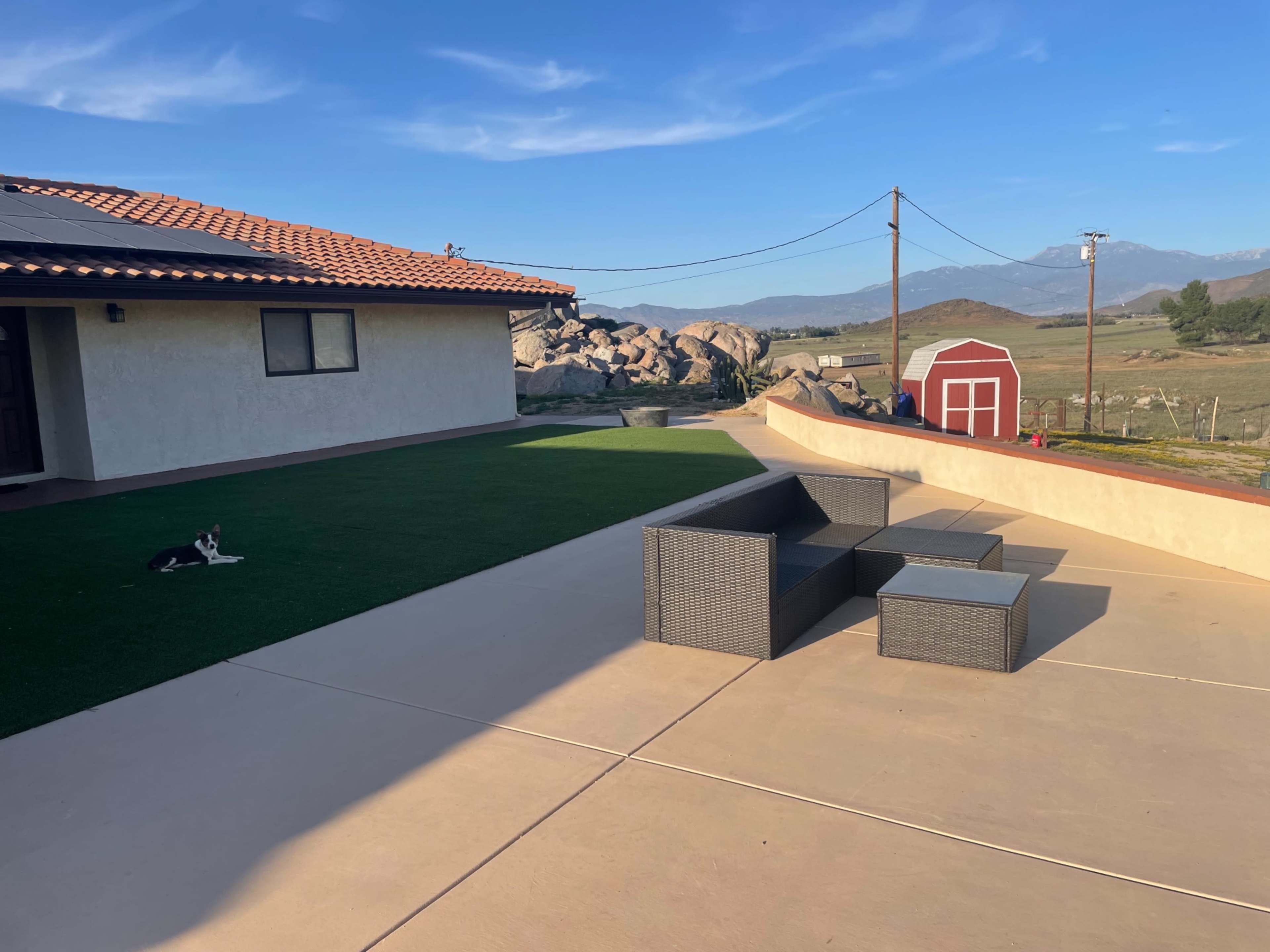 The scene features a patio area with a dark wicker seating set, a grassy section, and a red barn in the background against a mountain range.