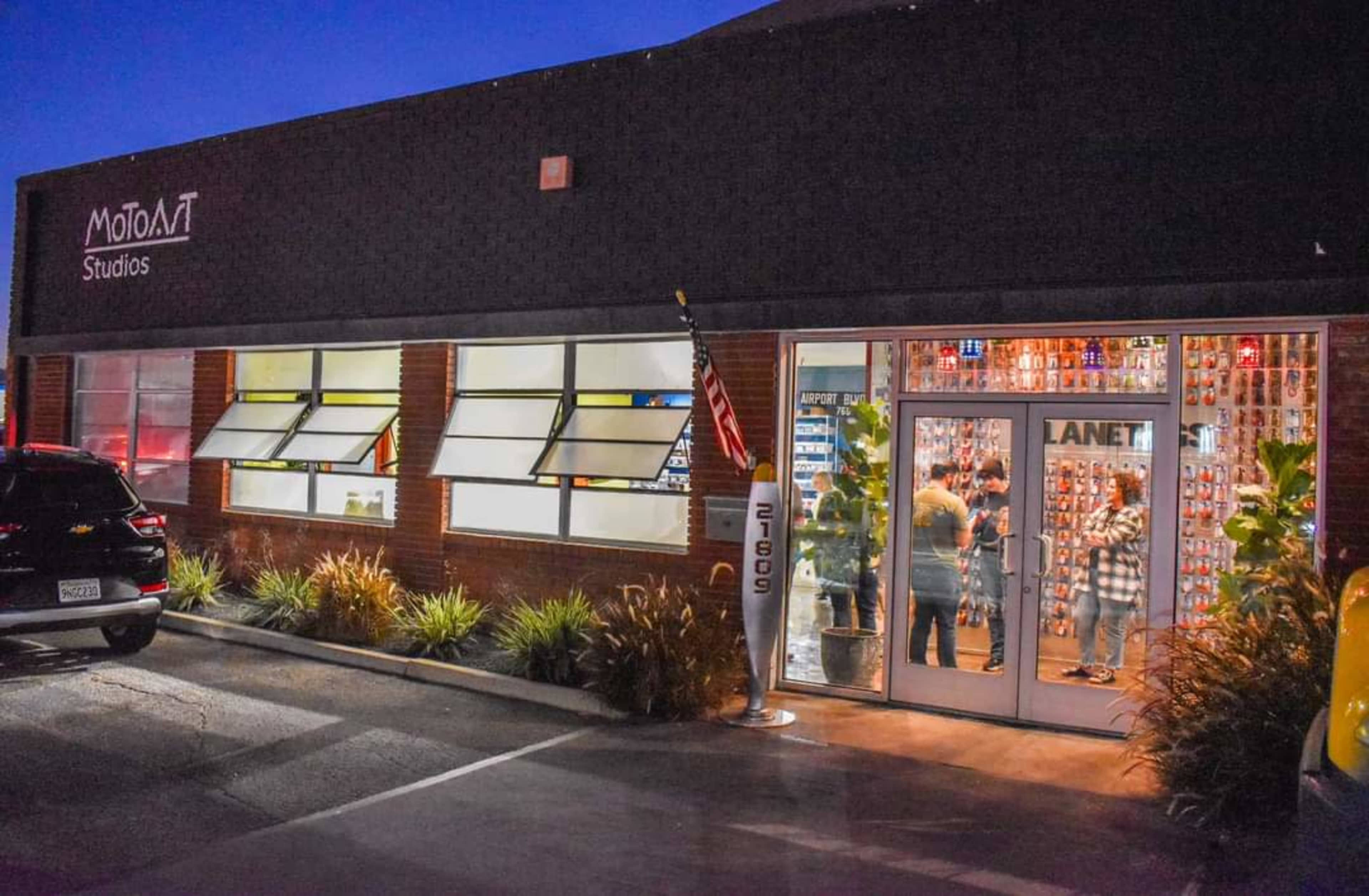 The image shows the exterior of MotoMat Studios at dusk, featuring a glass entrance with people visible inside and a U.S. flag displayed outside.