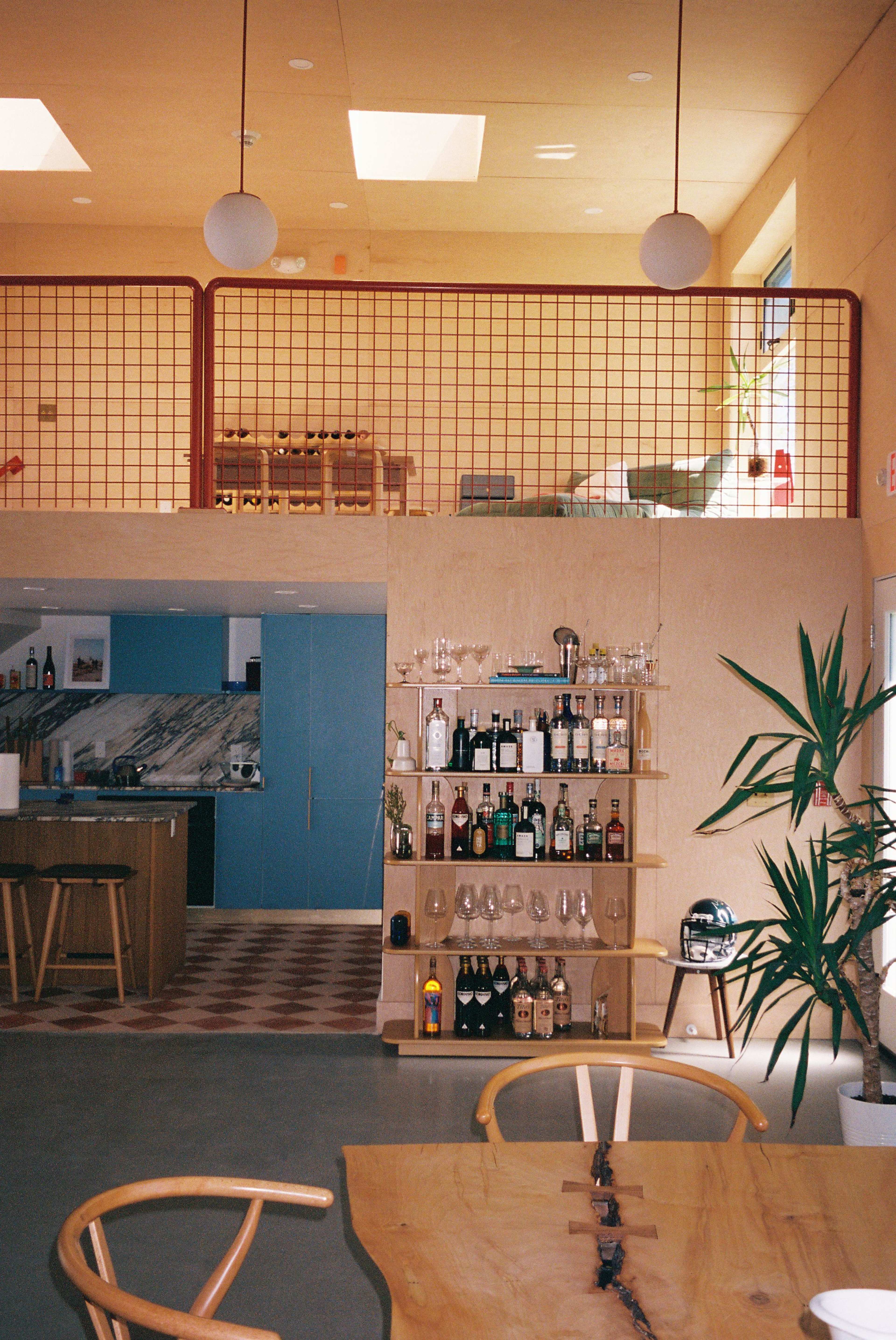 The image shows a modern interior design of a spacious room with a wooden bar, displays of liquor bottles, and a staircase leading to an upper level with additional seating and large windows.
