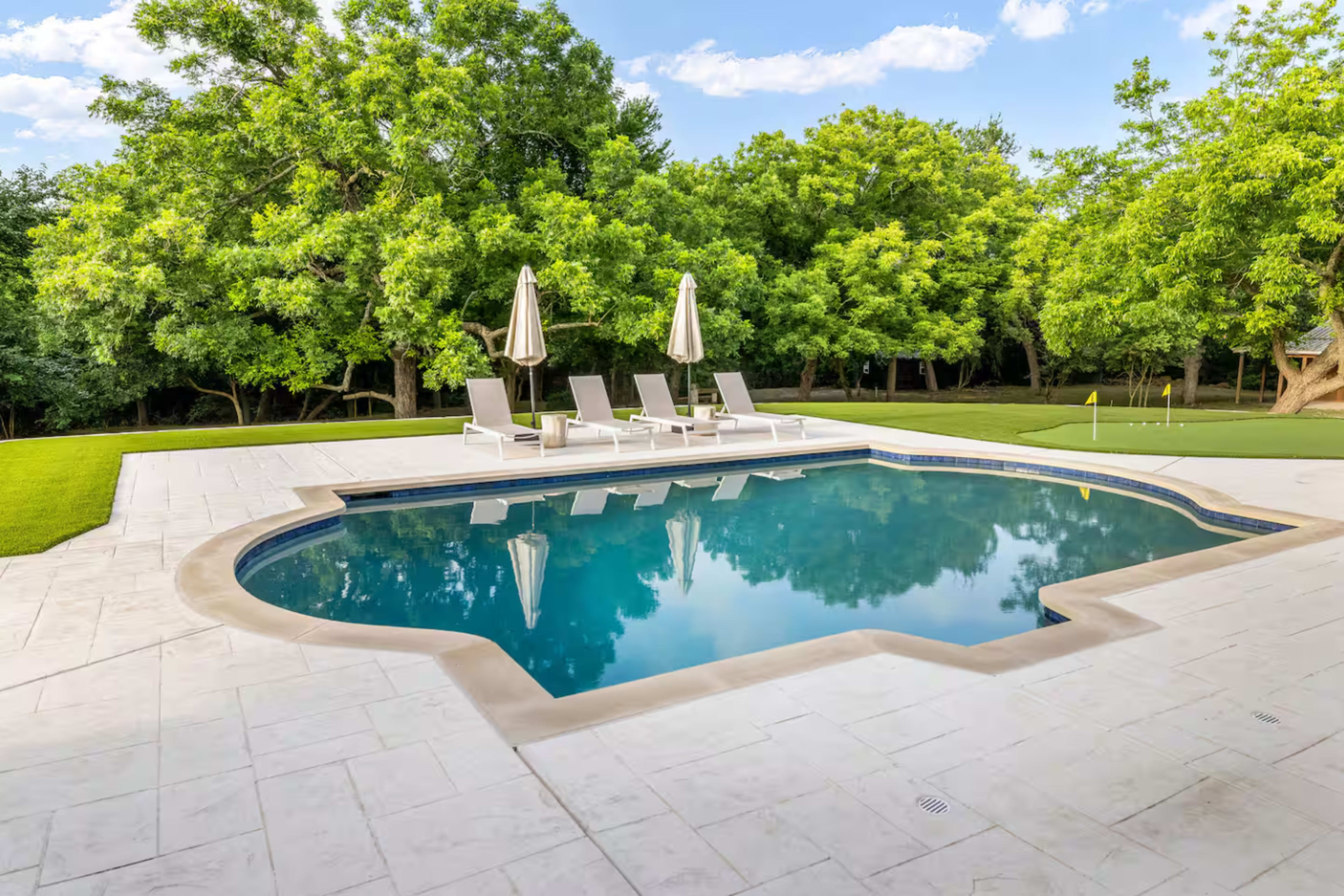 The image shows a swimming pool surrounded by lounge chairs under shade umbrellas, with lush greenery in the background.