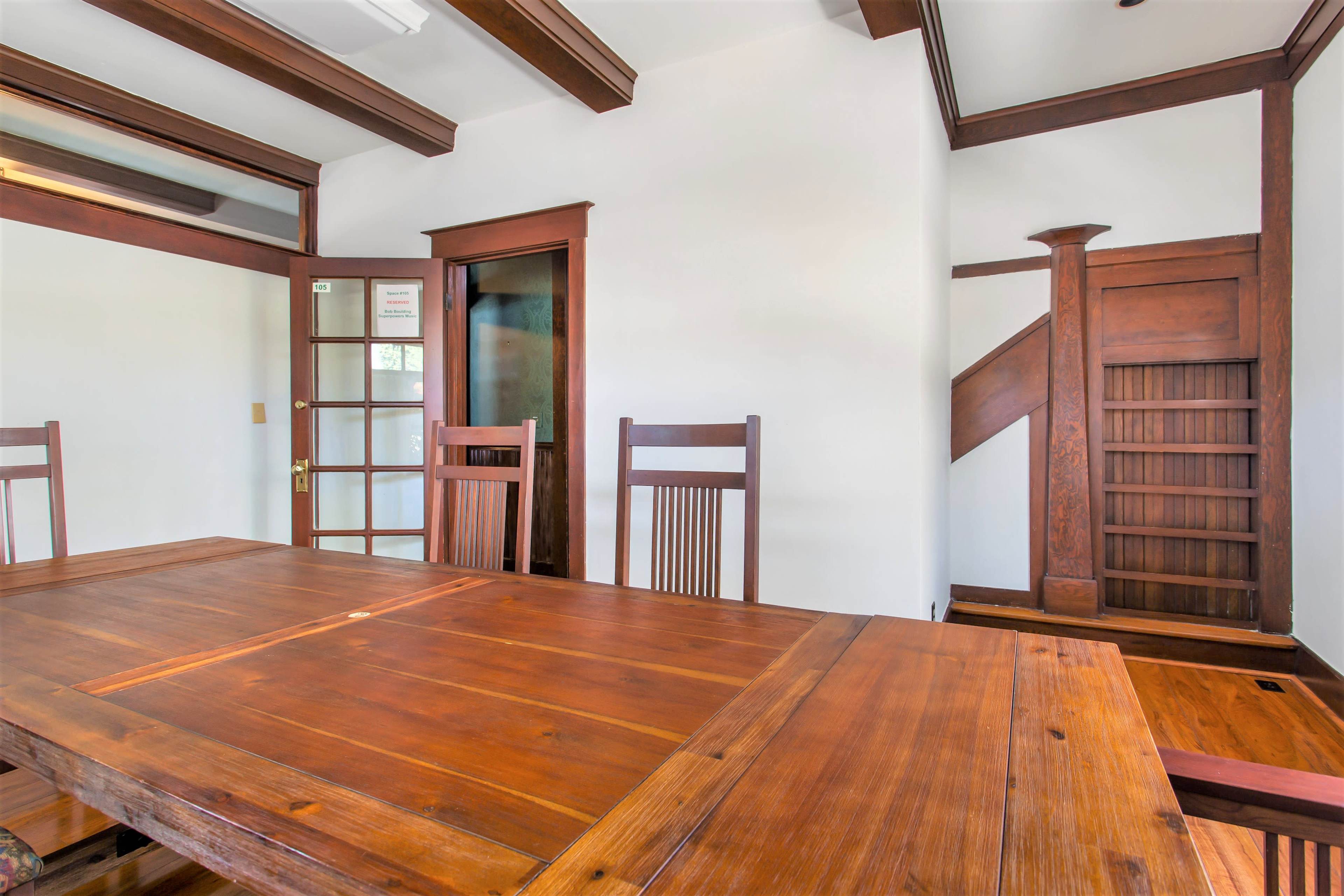 A wooden dining table with several chairs is positioned in a room featuring white walls and dark wooden beams, with a staircase and a glass-paneled door in the background.