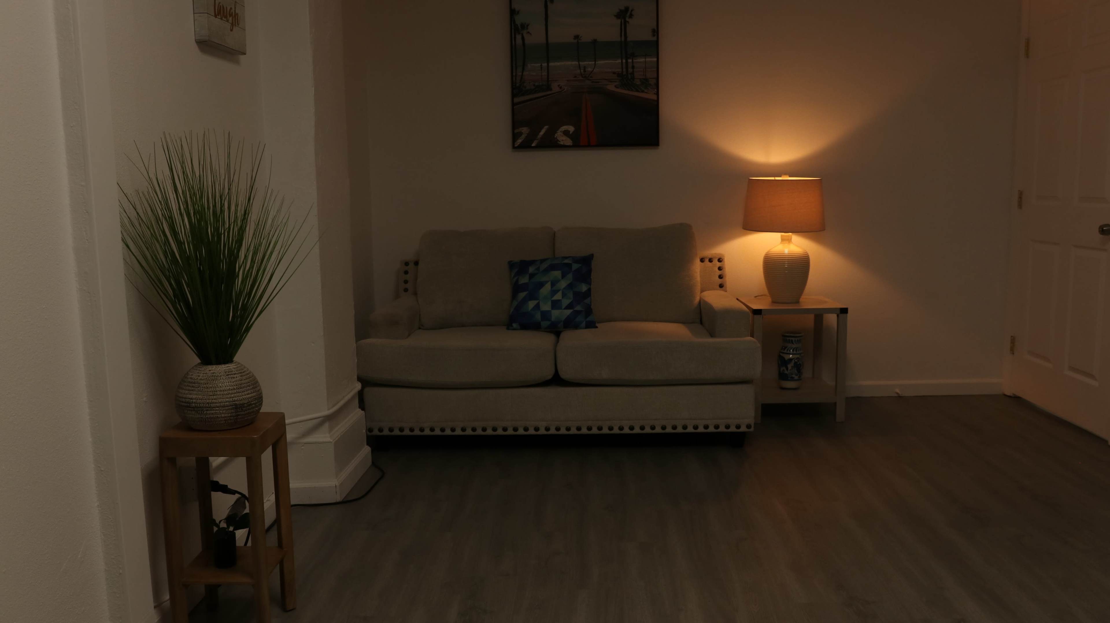 The image shows a softly lit living room featuring a beige couch with decorative pillows, a small side table with a lamp, and a plant in a pot against the wall.