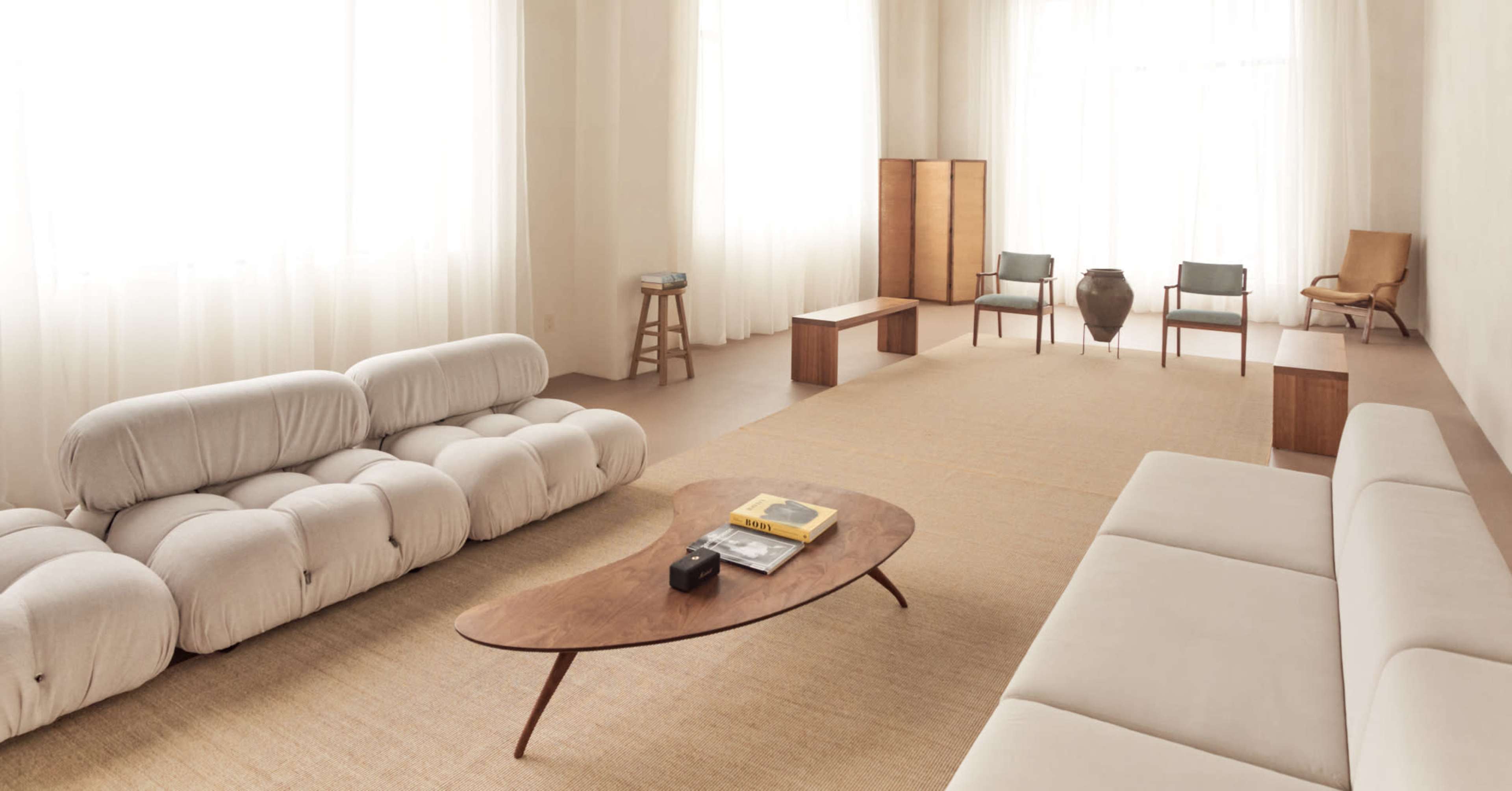 The image shows a spacious, minimalist living room with two sofas, a wooden coffee table, and several chairs arranged around a natural fiber rug under large windows with sheer curtains.