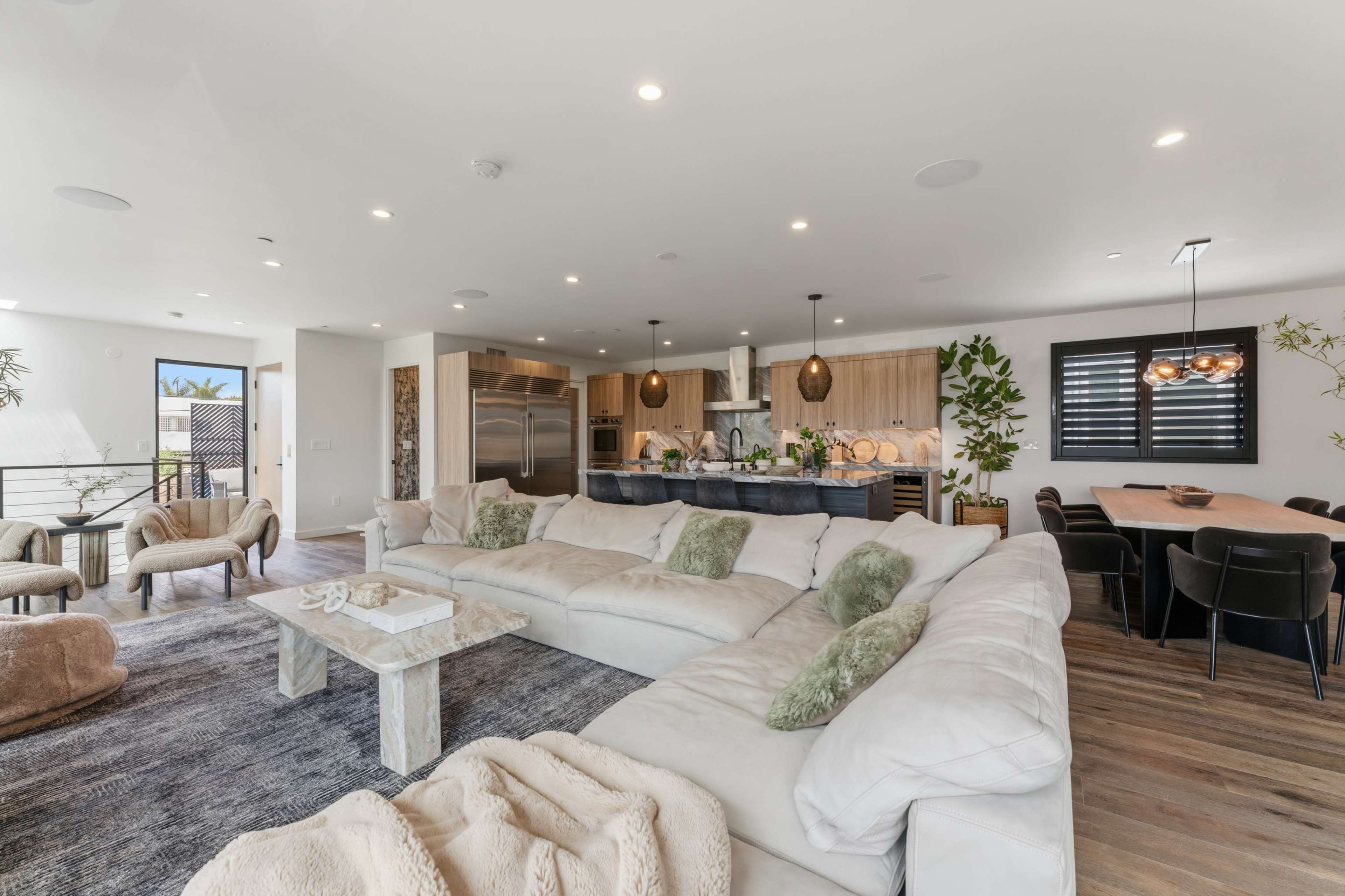The image shows a modern living room with a large sectional sofa, a dining area with a wooden table, and an open kitchen featuring sleek cabinetry and appliances.