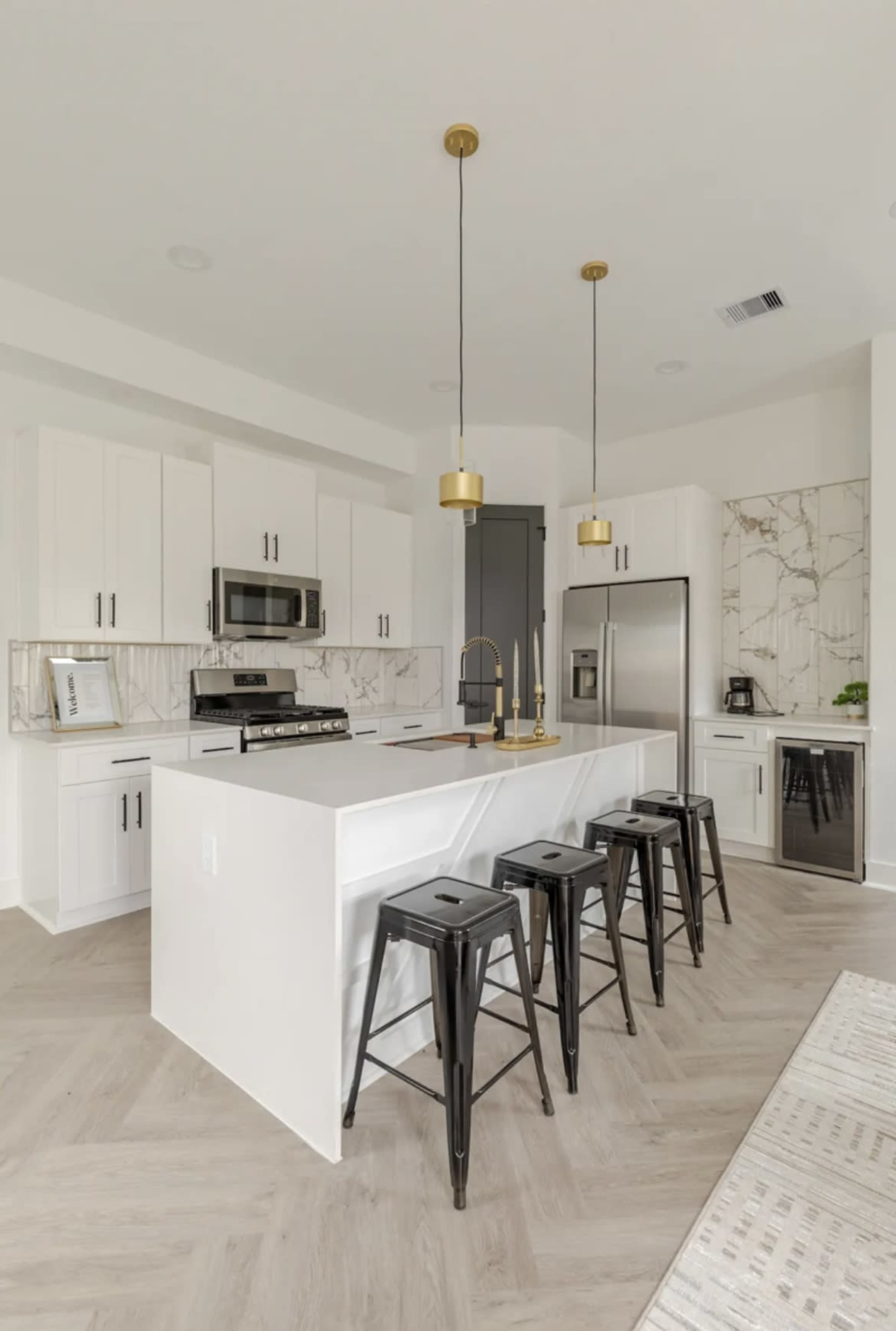 A modern kitchen features a light-colored island with black bar stools, stainless steel appliances, and marble backsplash accents.