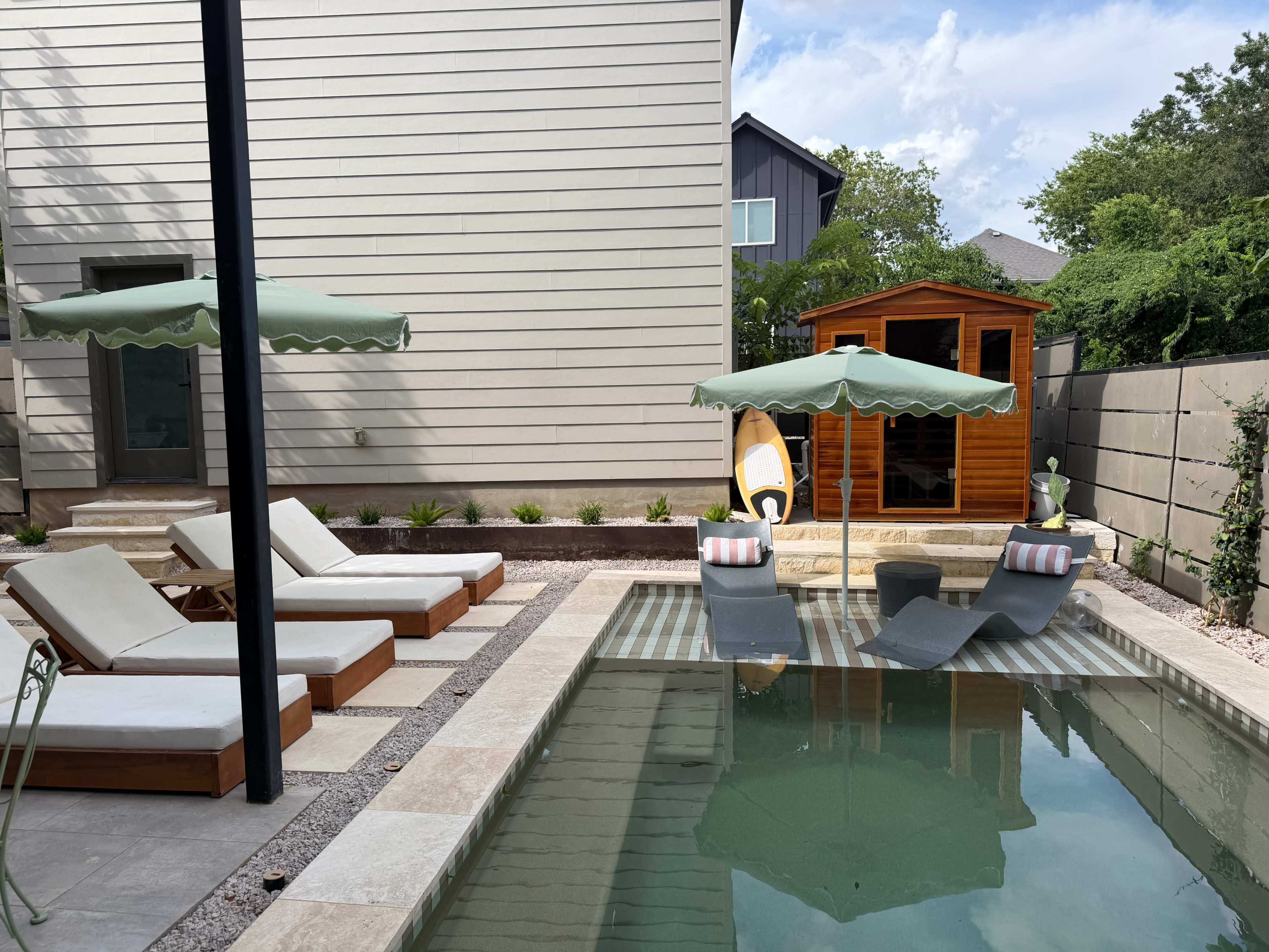 A rectangular swimming pool with two lounge chairs and an umbrella is situated beside a wooden shed in a residential backyard.
