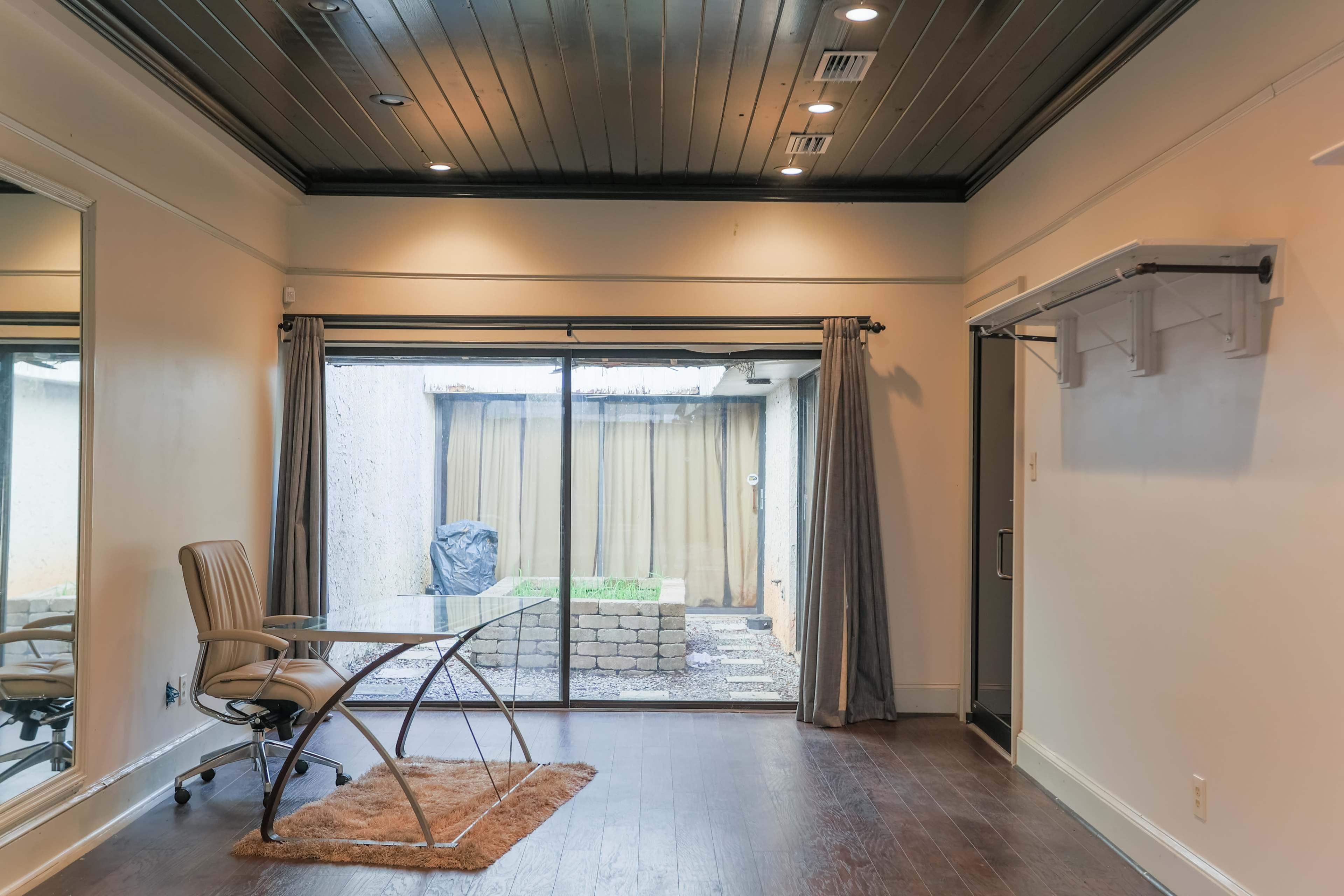 A room with a glass sliding door leading to a small outdoor area, a modern glass desk with a chair, and a large mirror on one wall.
