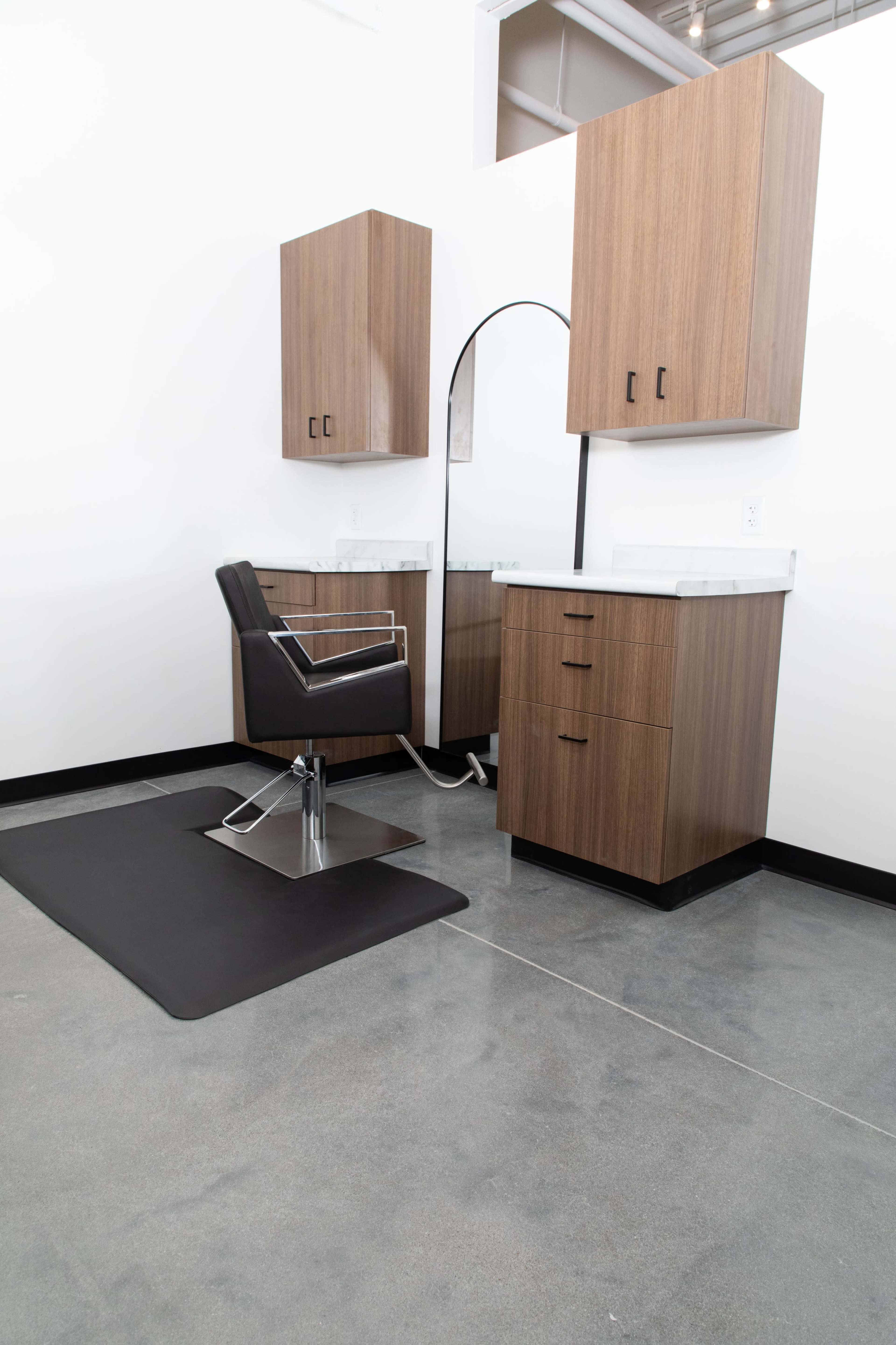 A modern hair salon setup features a black styling chair, a large mirror, and wooden cabinetry against a white wall.