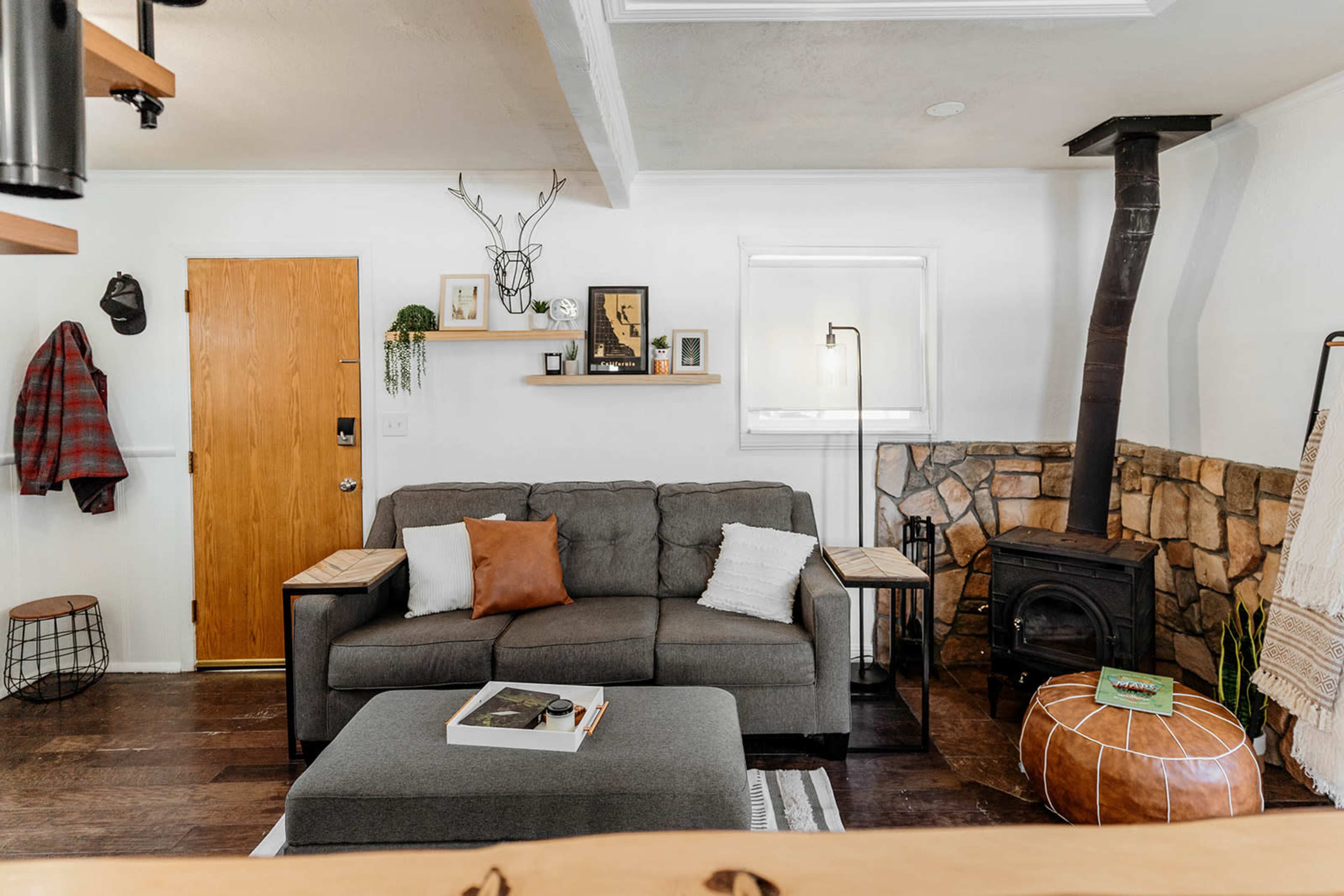 A living room featuring a gray sofa, a stone fireplace, wooden shelves with decor, and a round leather pouf.