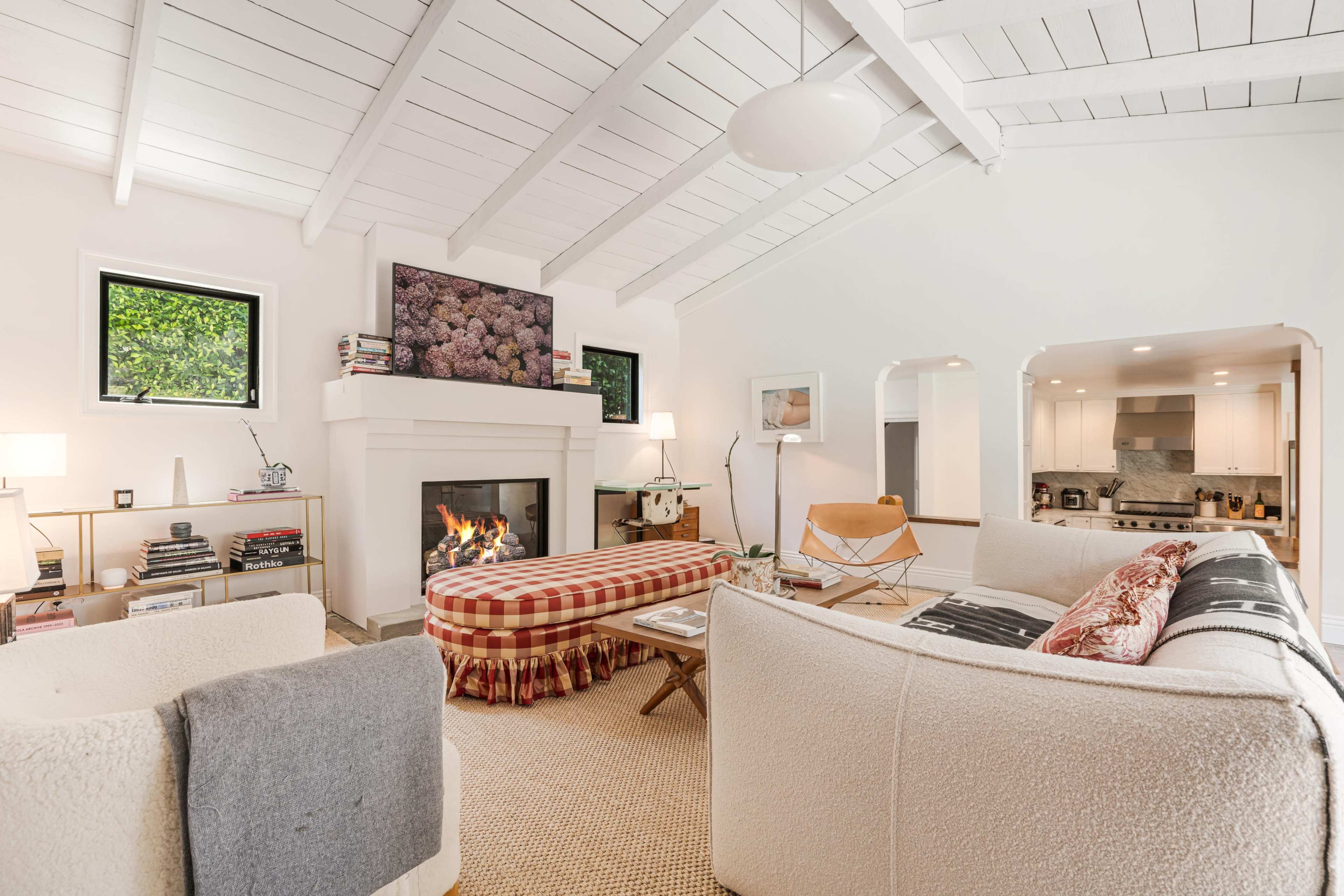A living room features a fireplace with a modern design, light-colored furnishings, a checkered ottoman, and large windows allowing natural light.