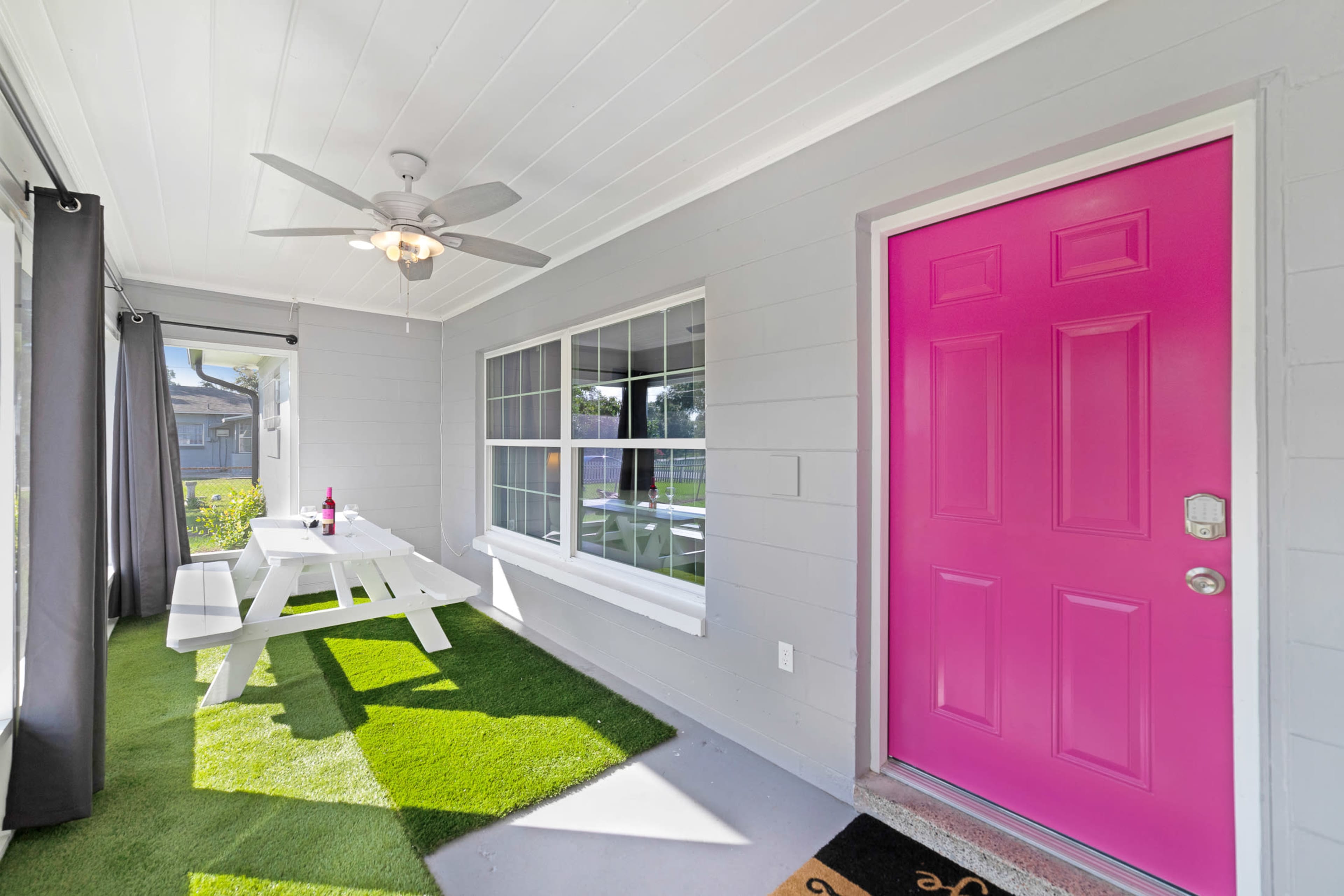 A bright pink door stands out against a light gray exterior, leading to a porch with a picnic table and artificial grass.