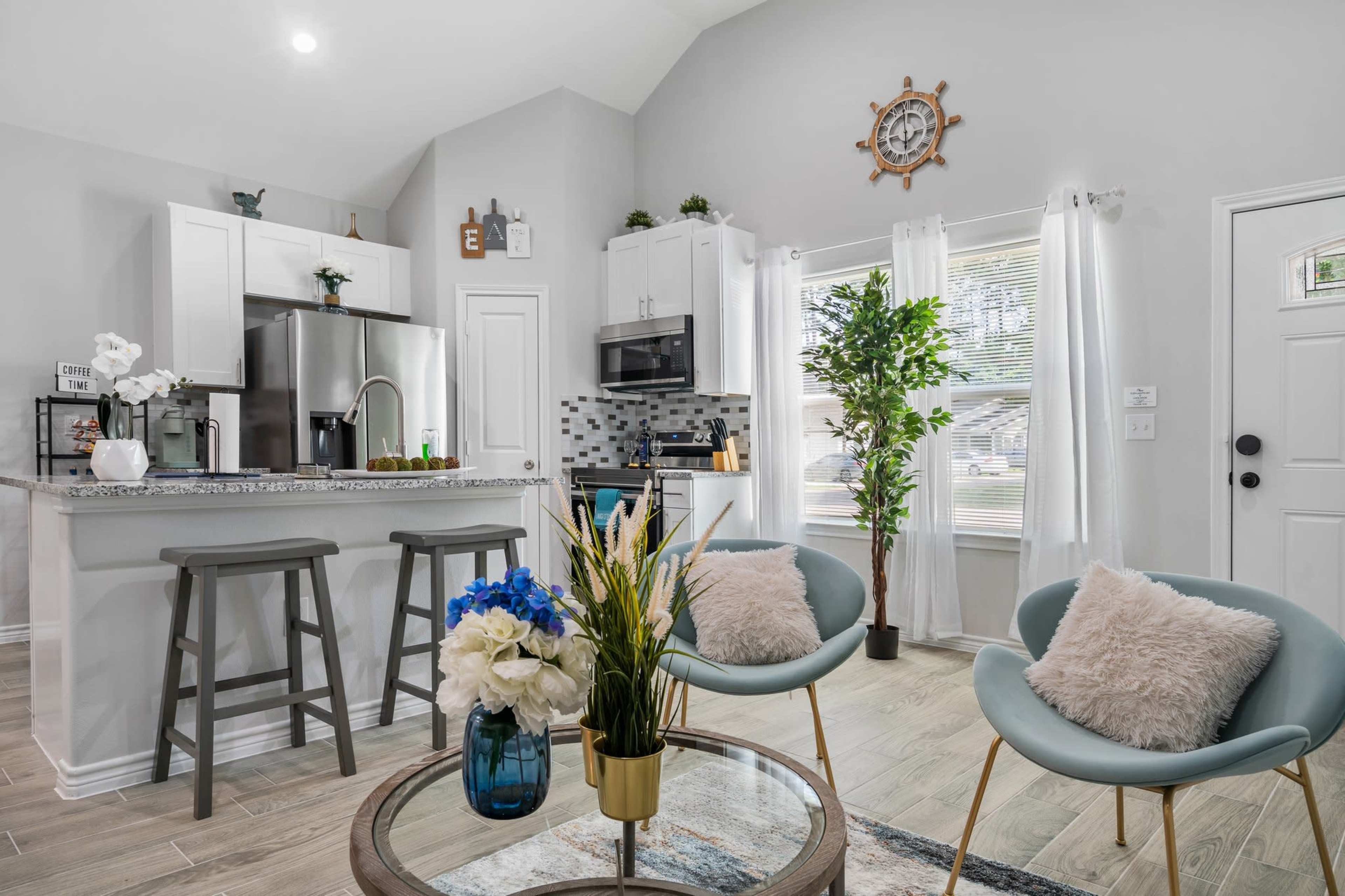 The image shows a modern kitchen and living area with stainless steel appliances, a circular coffee table, and two light blue chairs, surrounded by decorative plants and natural light.
