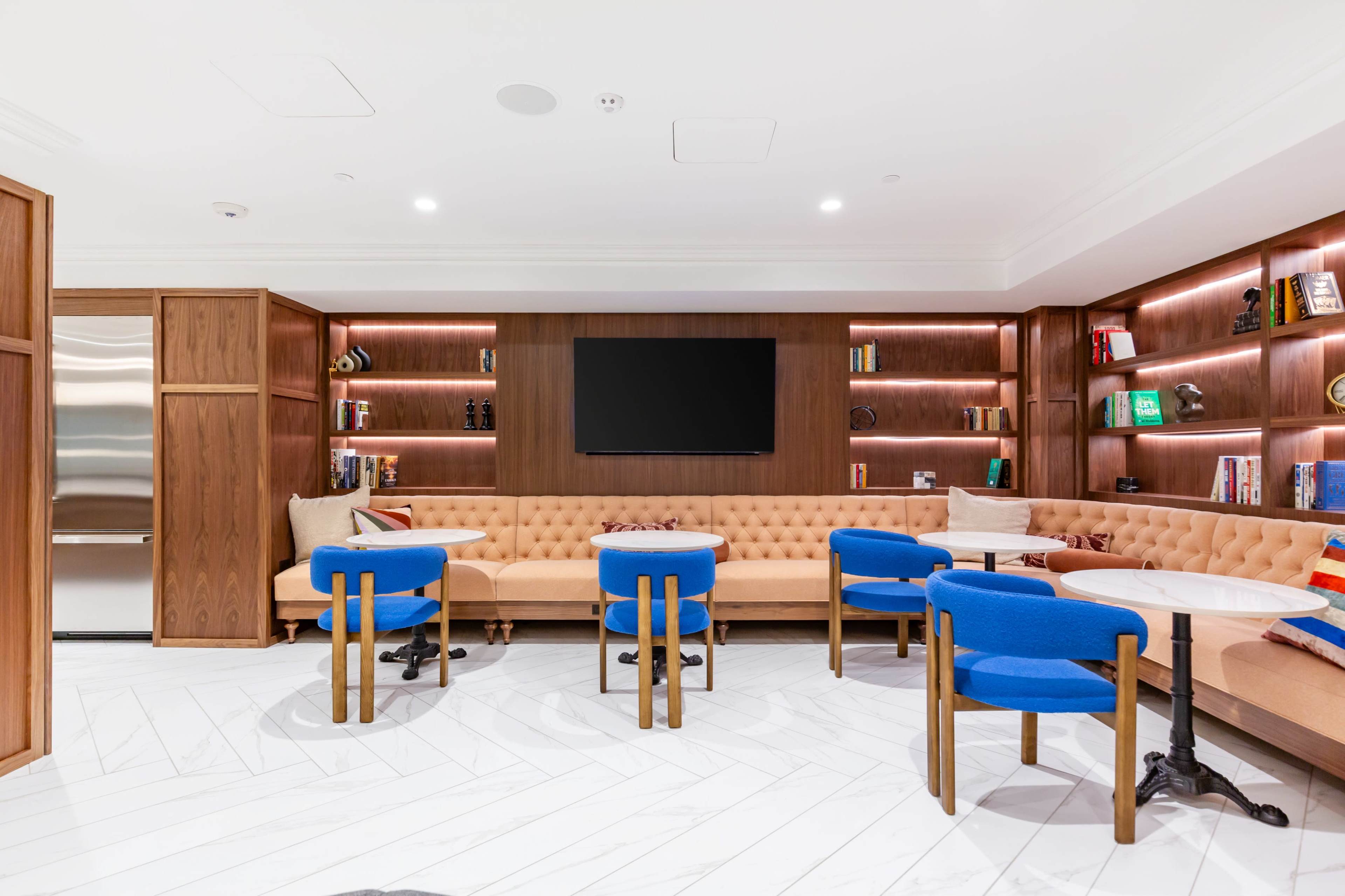 The image shows a cozy lounge area with a tufted beige couch, blue chairs, and bookshelves illuminated by warm lighting.