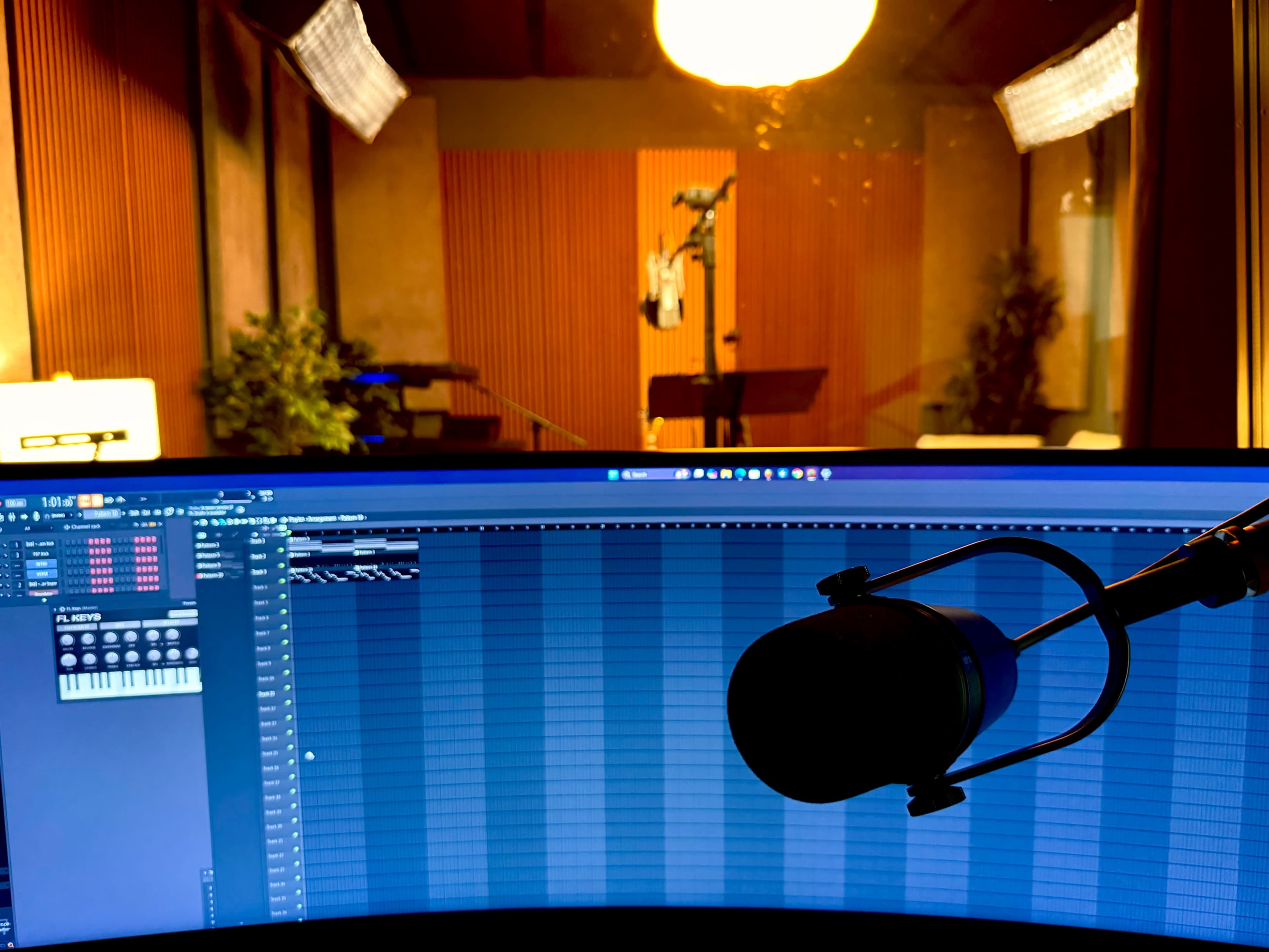 The image shows a recording studio with a microphone in the foreground and sound equipment visible on a computer screen, while soundproofed walls are seen in the background.