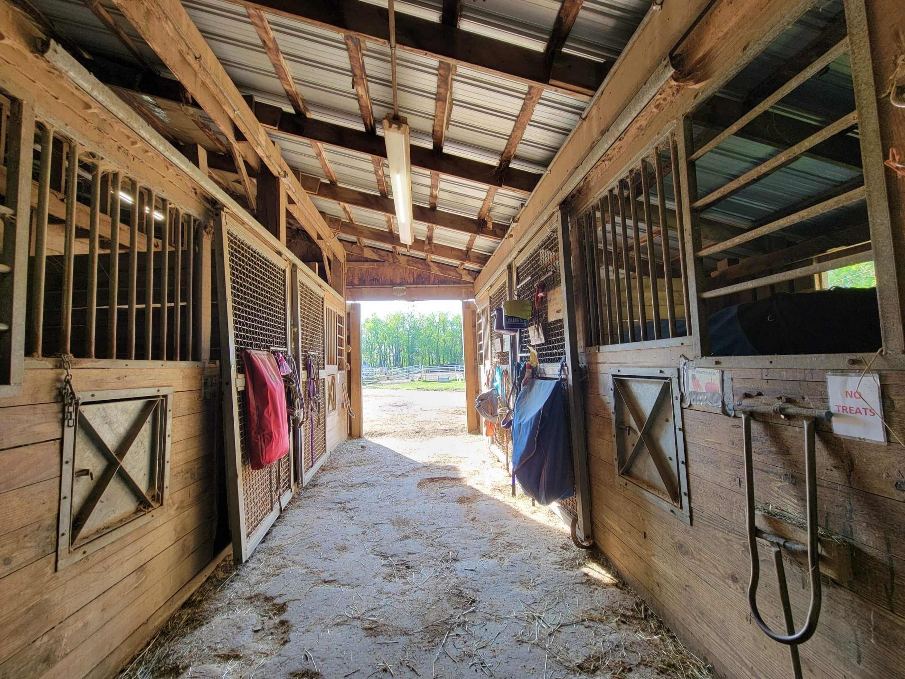 The interior of a horse barn with two open stalls on either side and a view of an outdoor pasture at the end of the aisle.