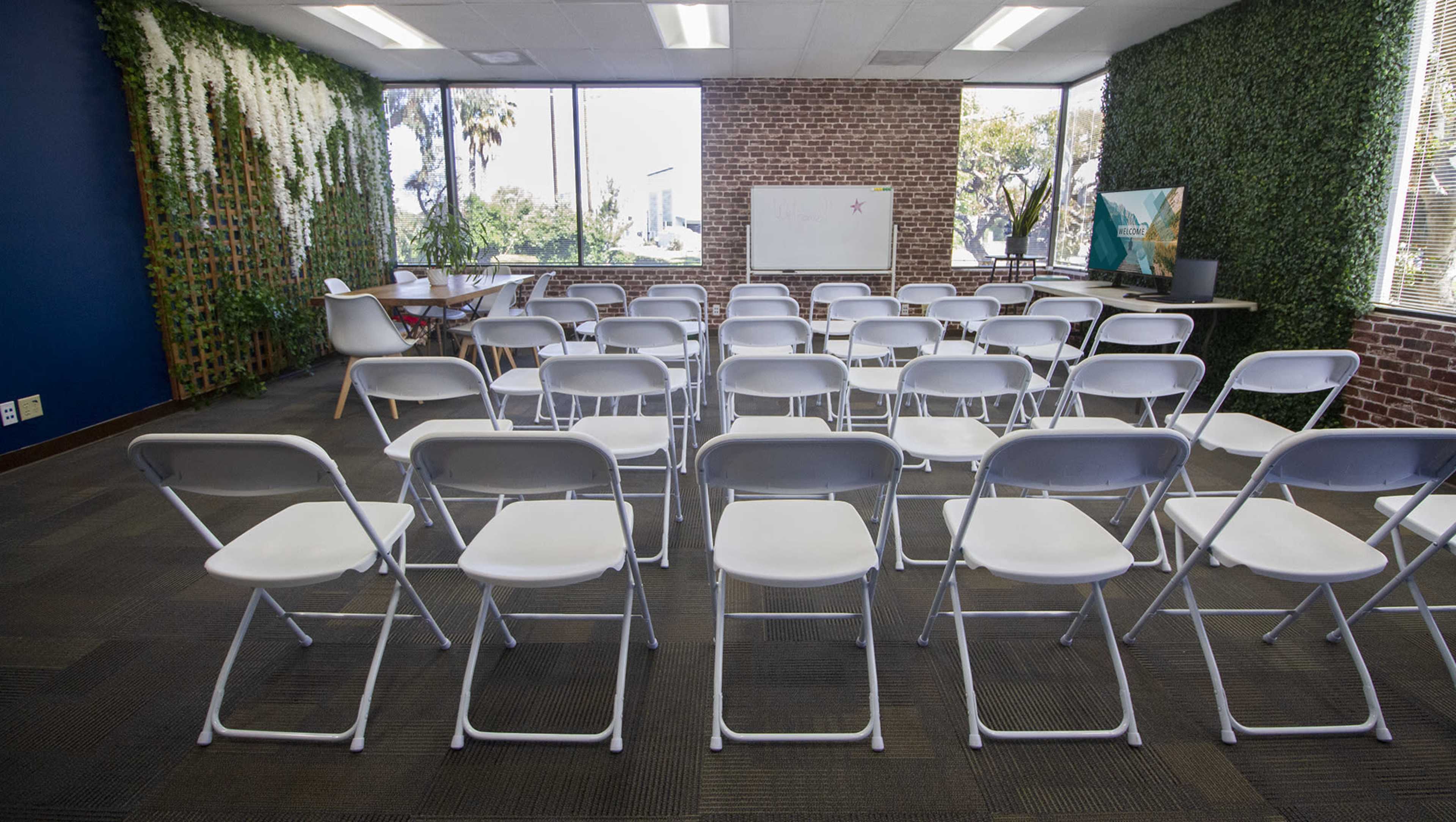 The image shows a seminar room arranged with rows of white folding chairs facing a blank whiteboard, surrounded by two large windows and greenery on the walls.