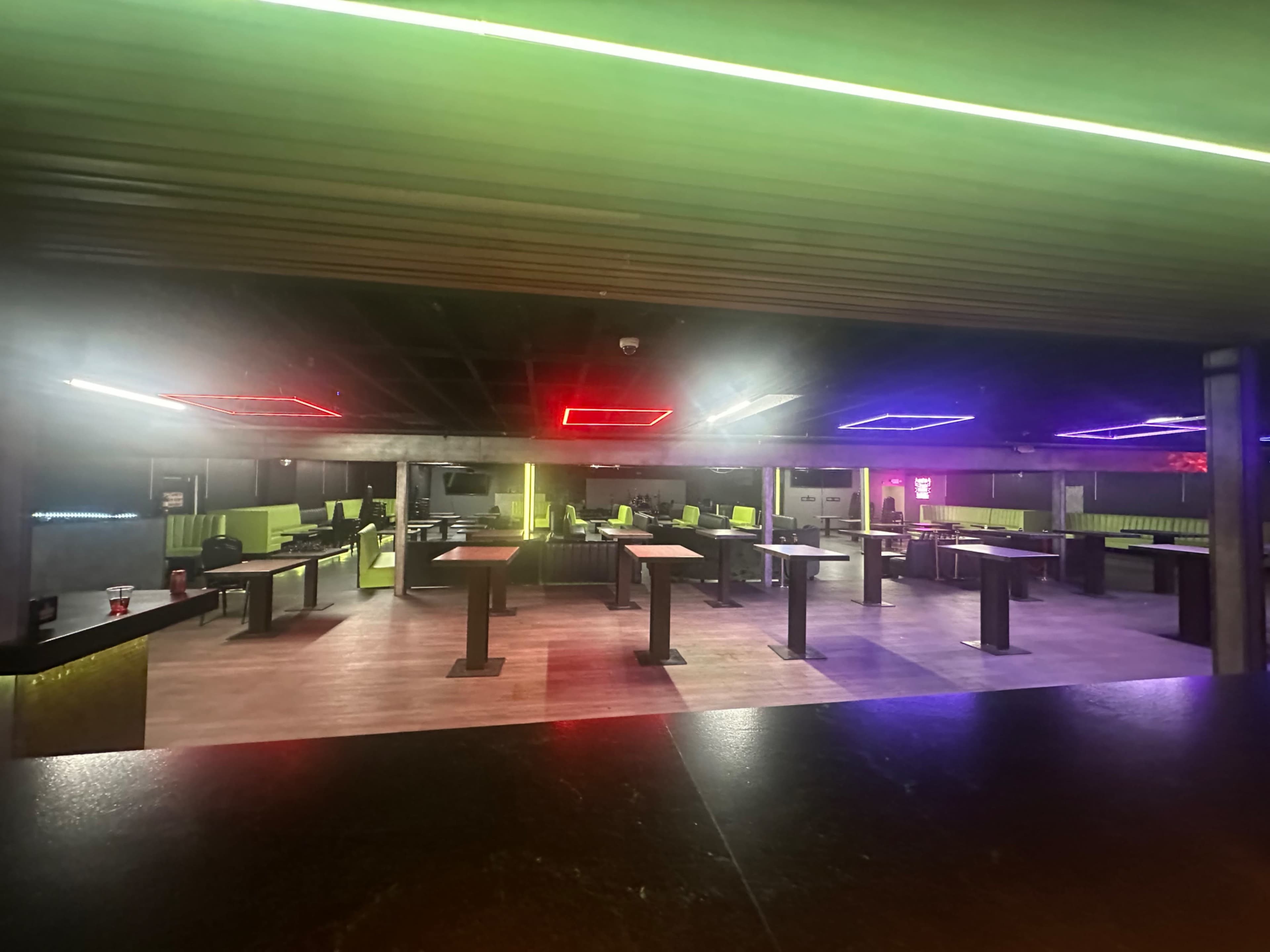 The image shows a dimly lit, modern indoor space with green and purple neon lights, featuring several tables and chairs arranged in rows.