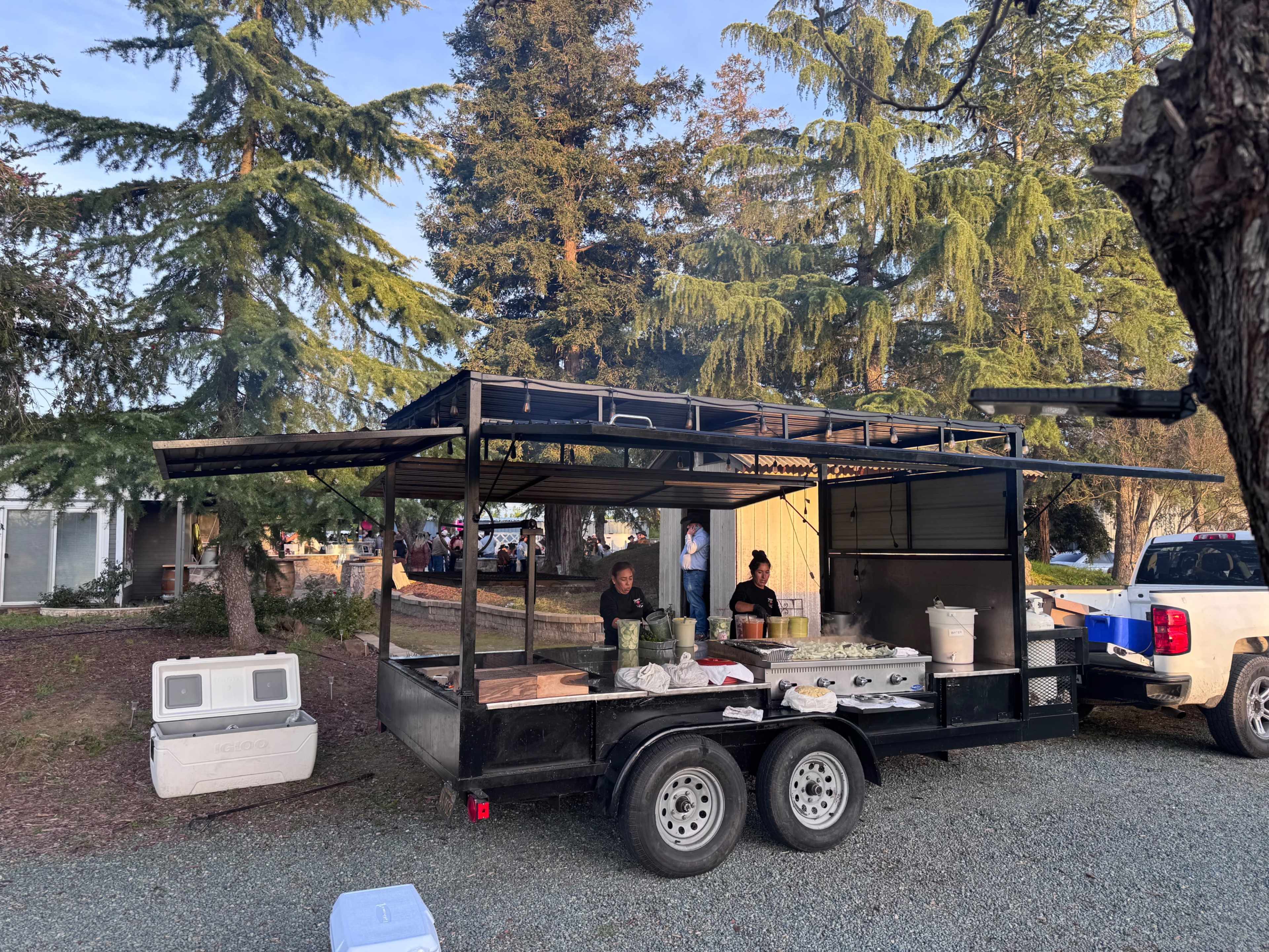 A food truck is set up in a gravel area, with two people working inside and large trees in the background.