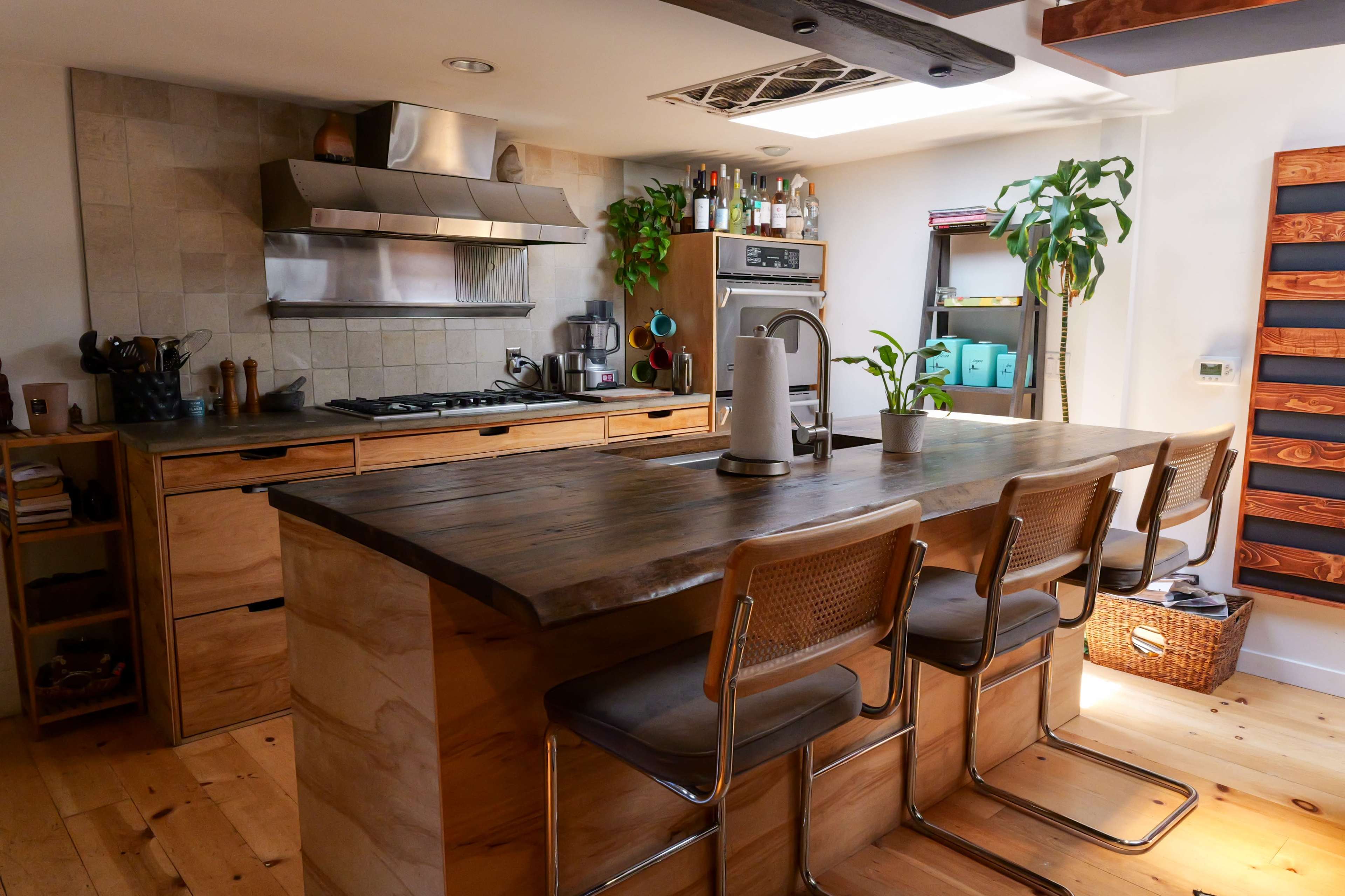 A modern kitchen features a wooden island with bar stools, stainless steel appliances, and potted plants in a well-lit space.