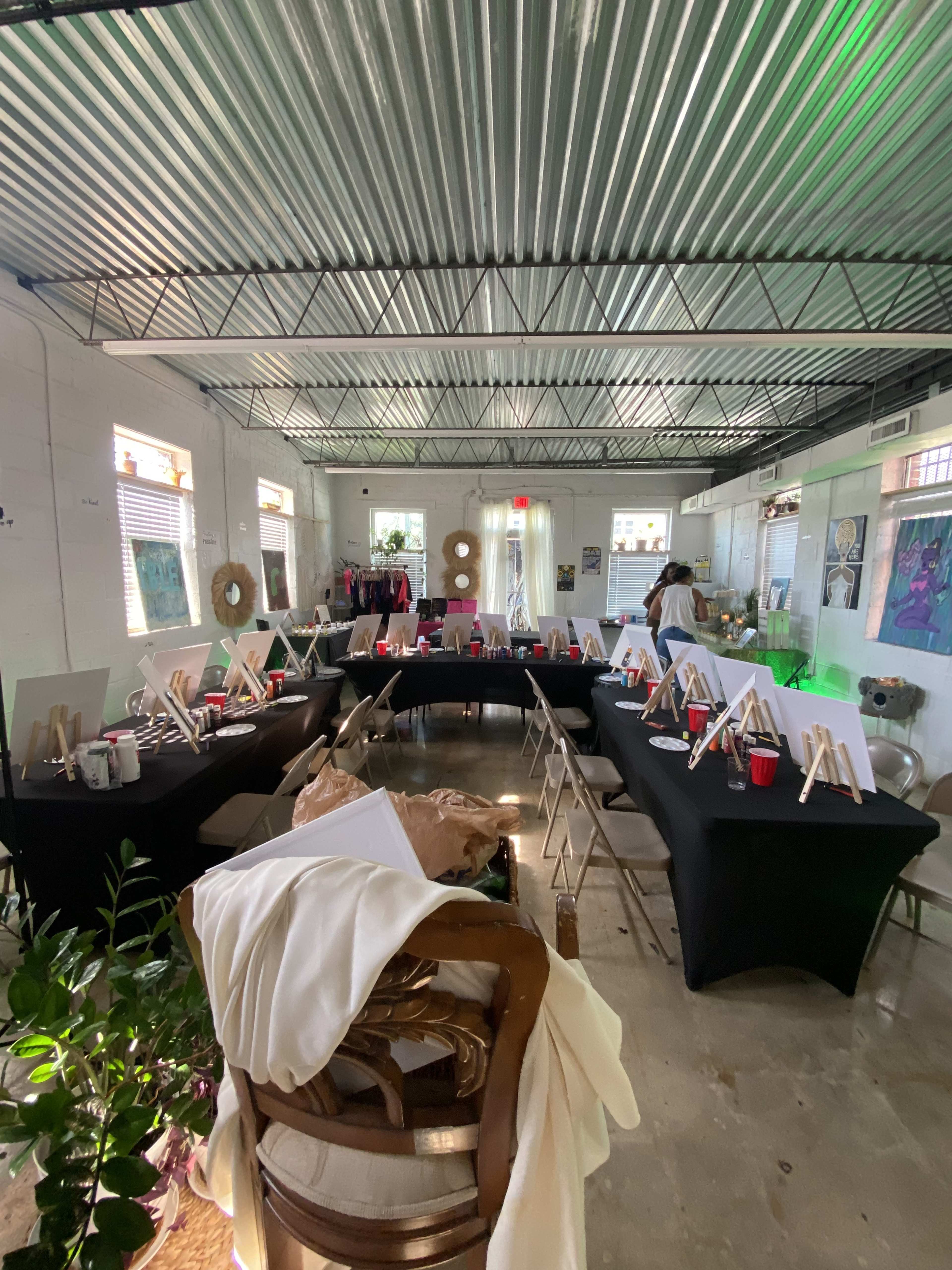 A spacious studio is set up for an art event, featuring multiple tables covered with black cloth, each displaying easels, canvases, and supplies.