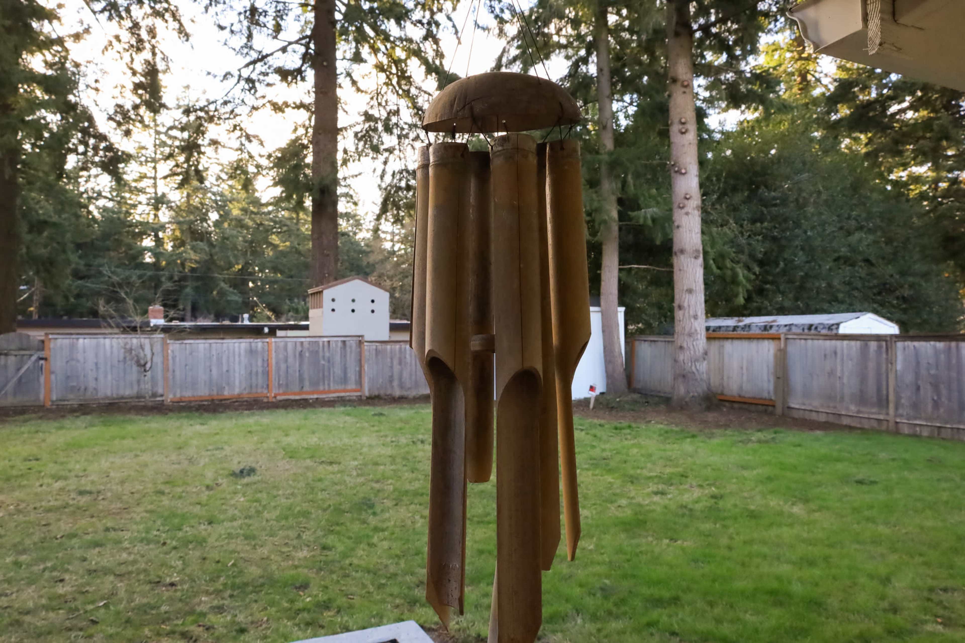 A wind chime made of wooden tubes hangs in a backyard surrounded by tall trees and a wooden fence.