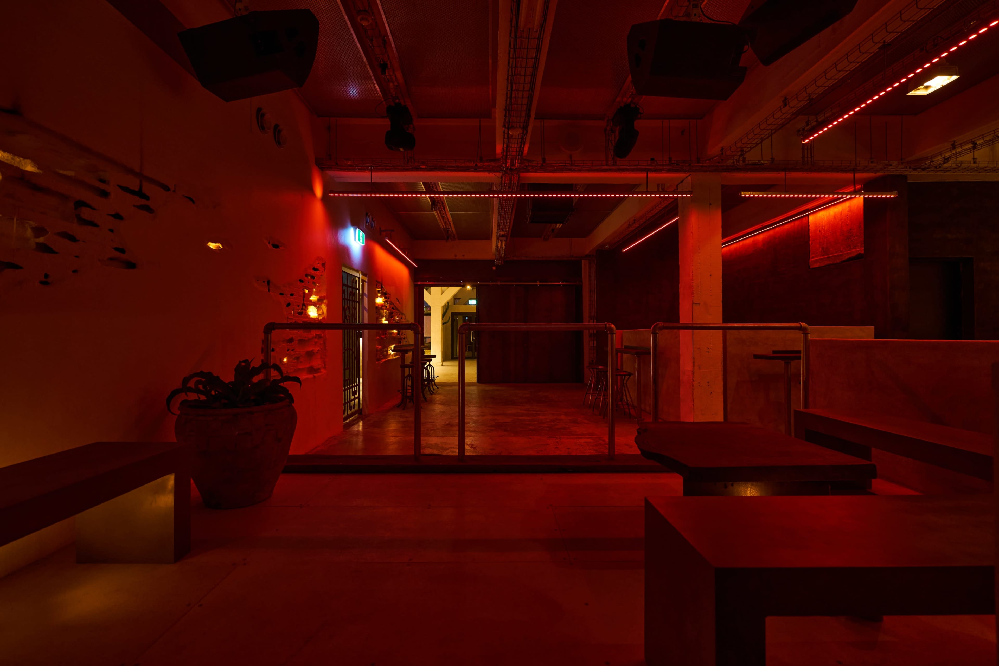 The image shows a dimly lit venue featuring industrial-style furniture with red lighting and a glass barrier separating two areas.
