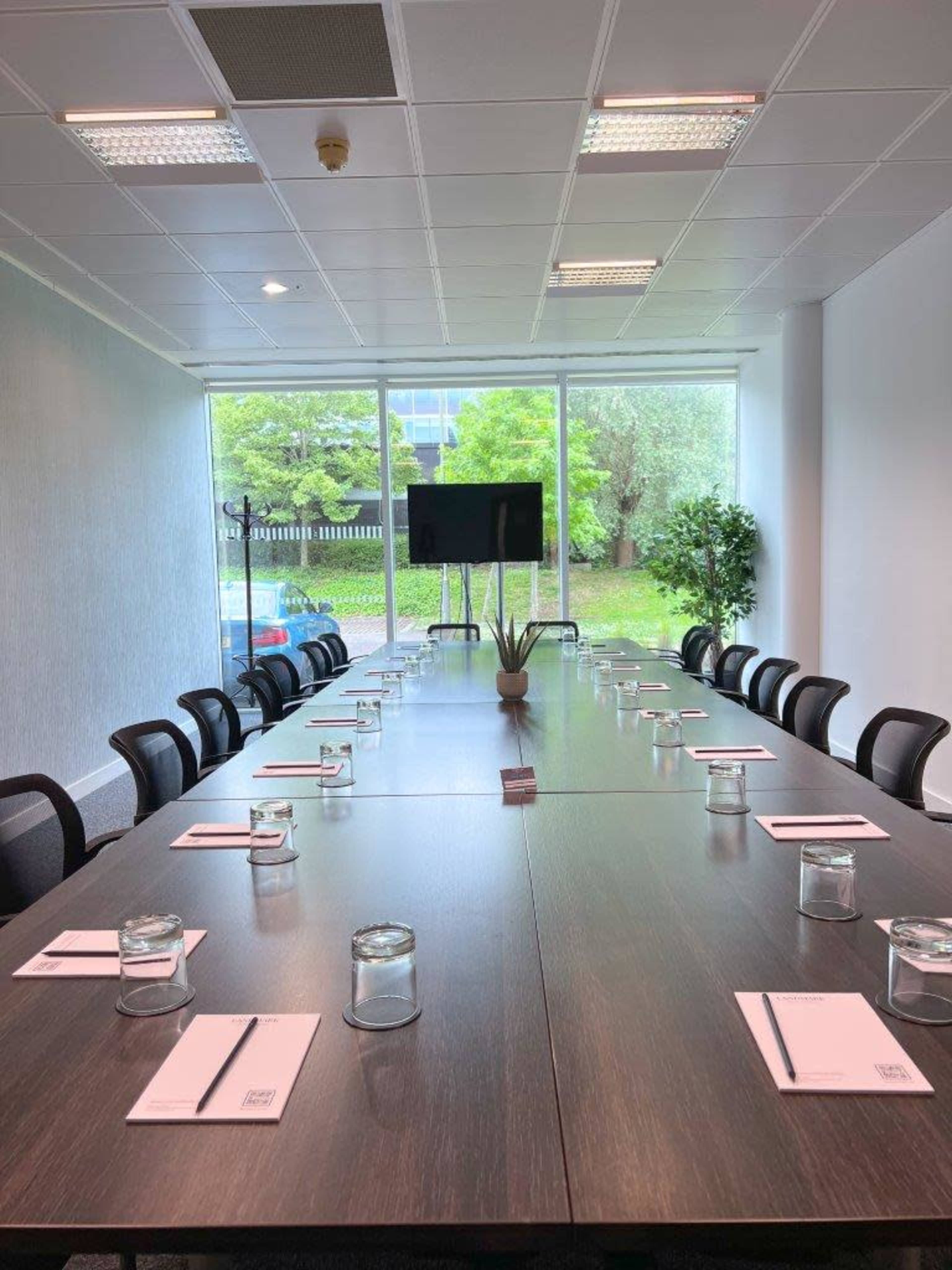 A long conference table is set with notepads and glass jars, positioned in a bright room with large windows overlooking greenery.