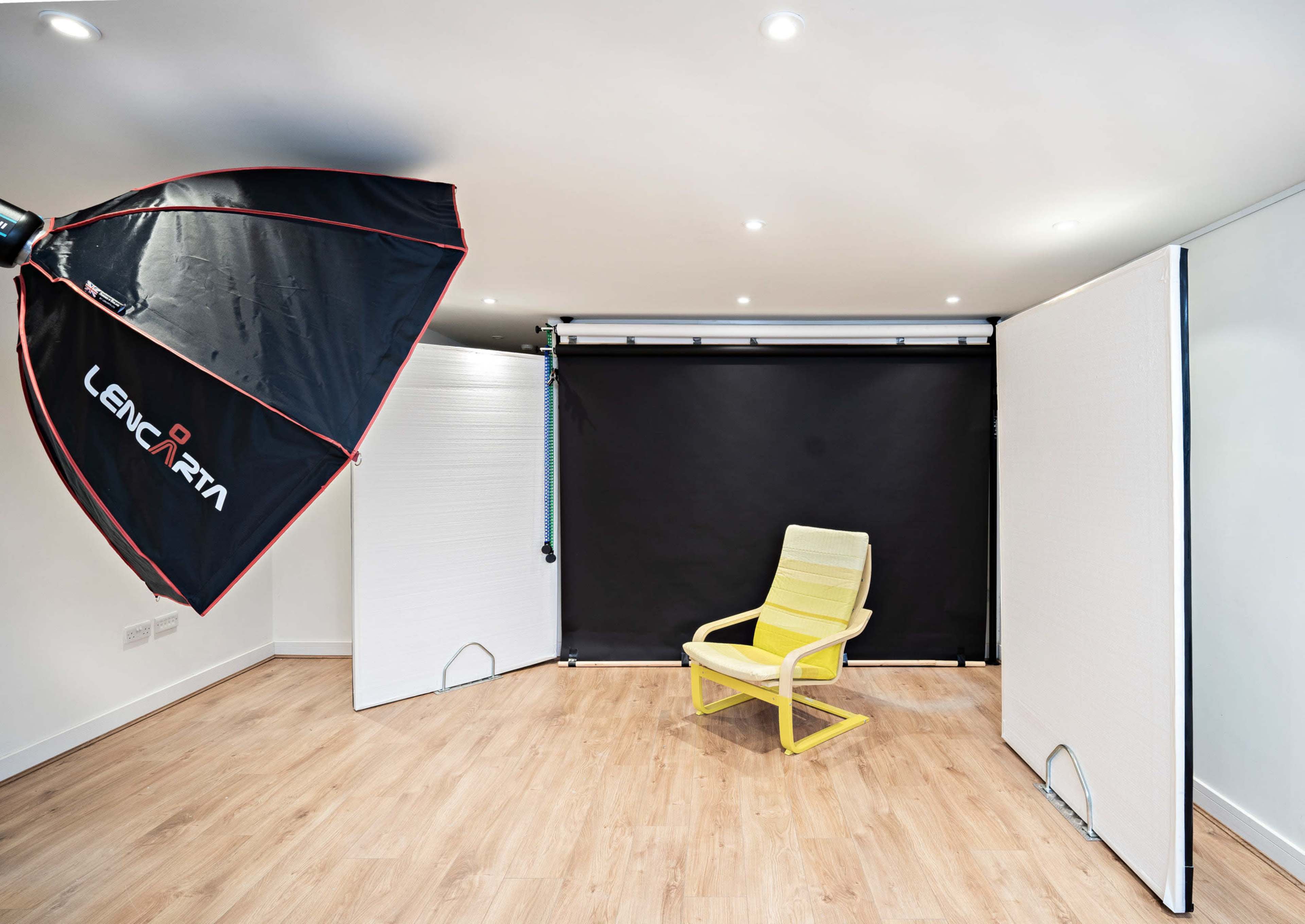 A yellow chair is positioned under a large softbox light in a minimalist photographic studio with black and white backdrop panels.