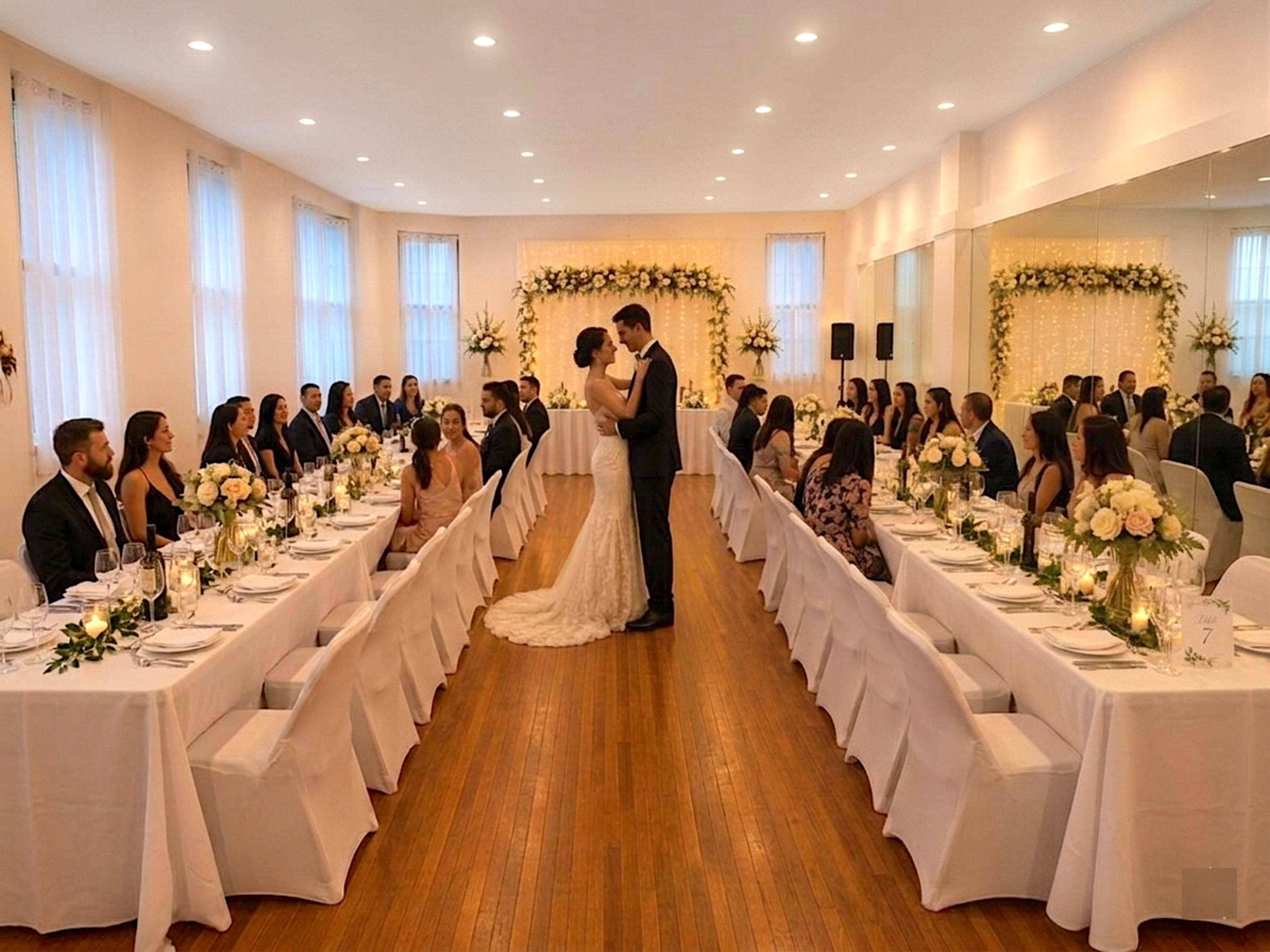 A couple is exchanging vows at a wedding ceremony in a decorated hall, surrounded by seated guests at long dining tables.