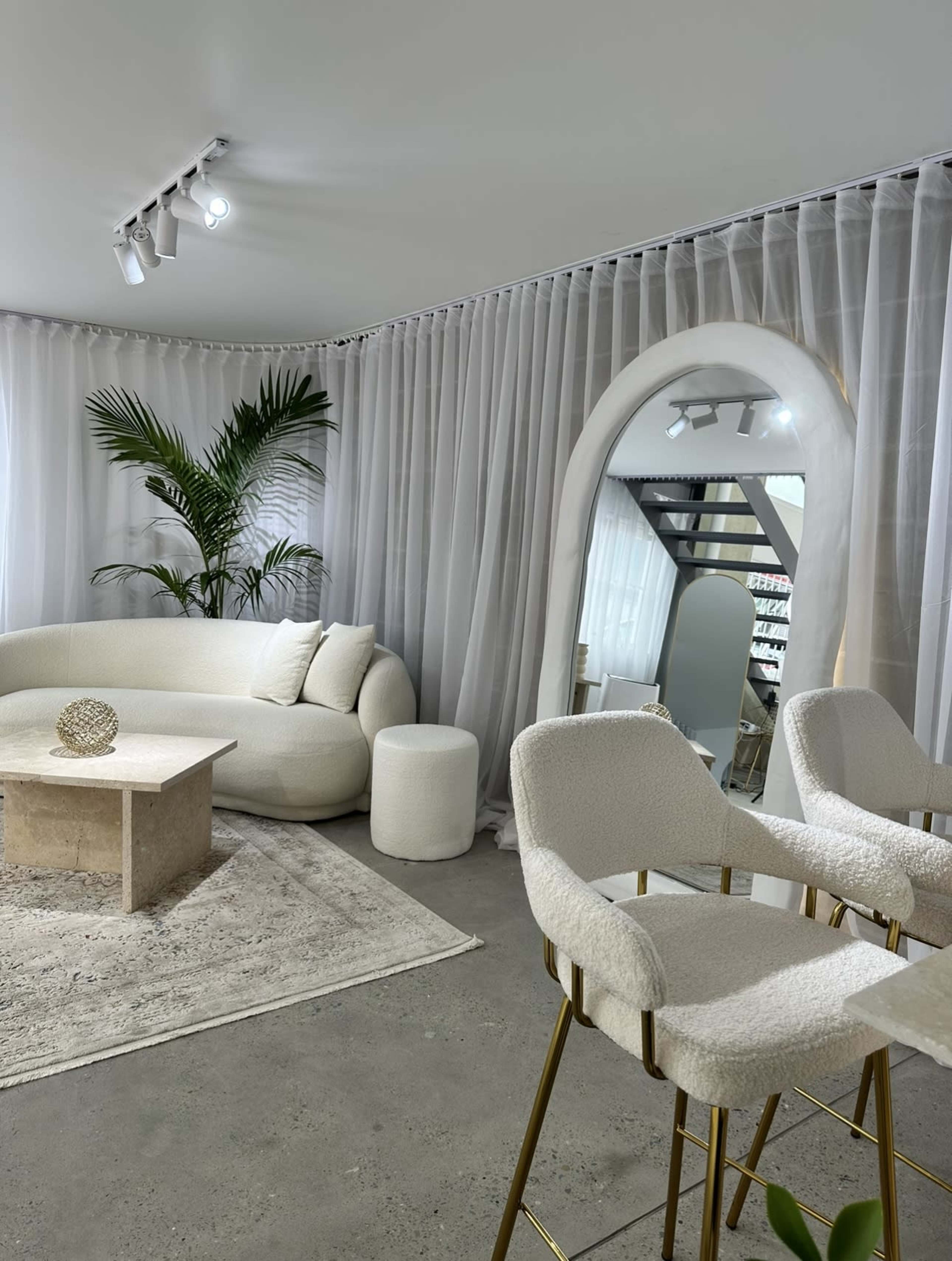 A modern interior space featuring a light-colored couch, a round seating stool, a stone coffee table, and a large mirror surrounded by sheer curtains and greenery.