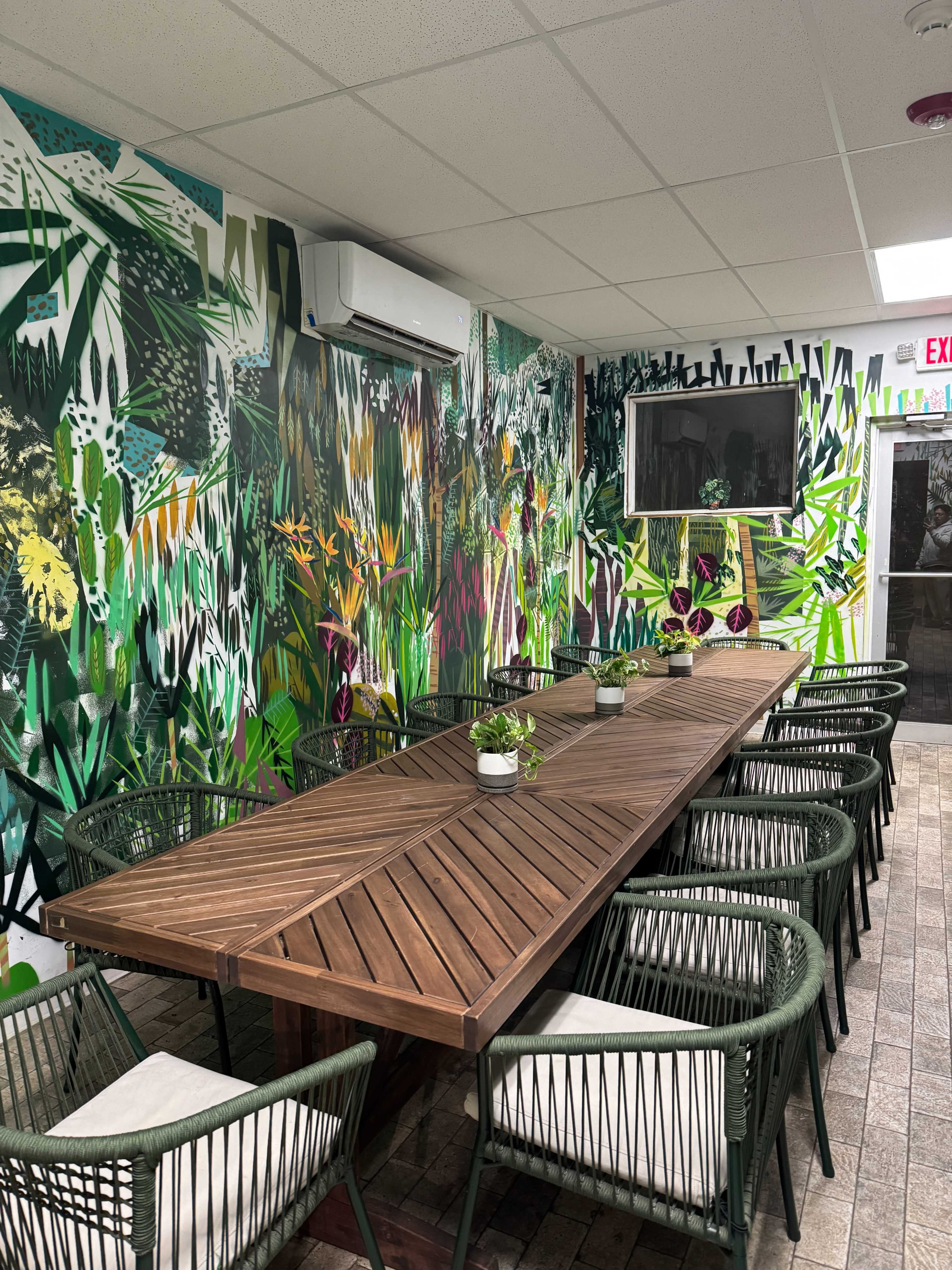 The image shows a dining area with a long wooden table surrounded by black wicker chairs, set against a colorful, botanical-themed mural on the walls.