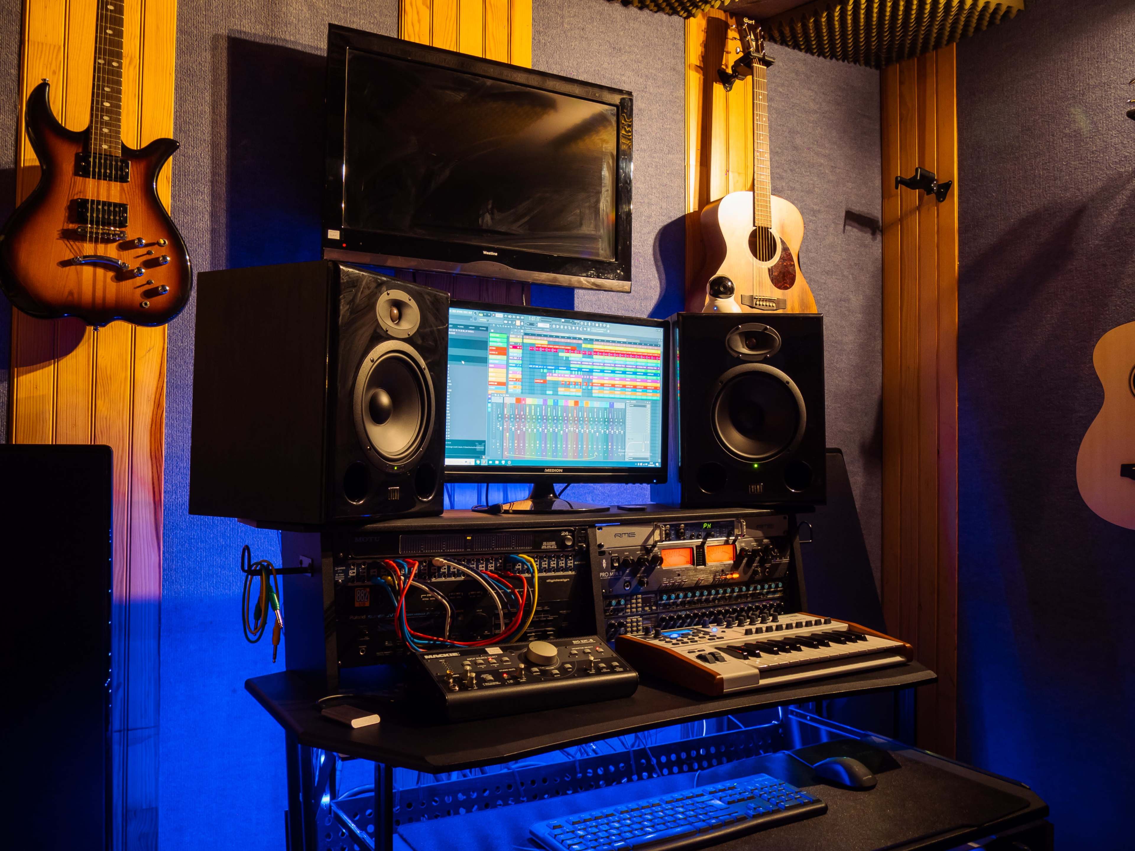 A music studio setup features a computer monitor displaying a digital audio workstation, flanked by speakers, a keyboard, and audio equipment, with guitars hanging on the wall.
