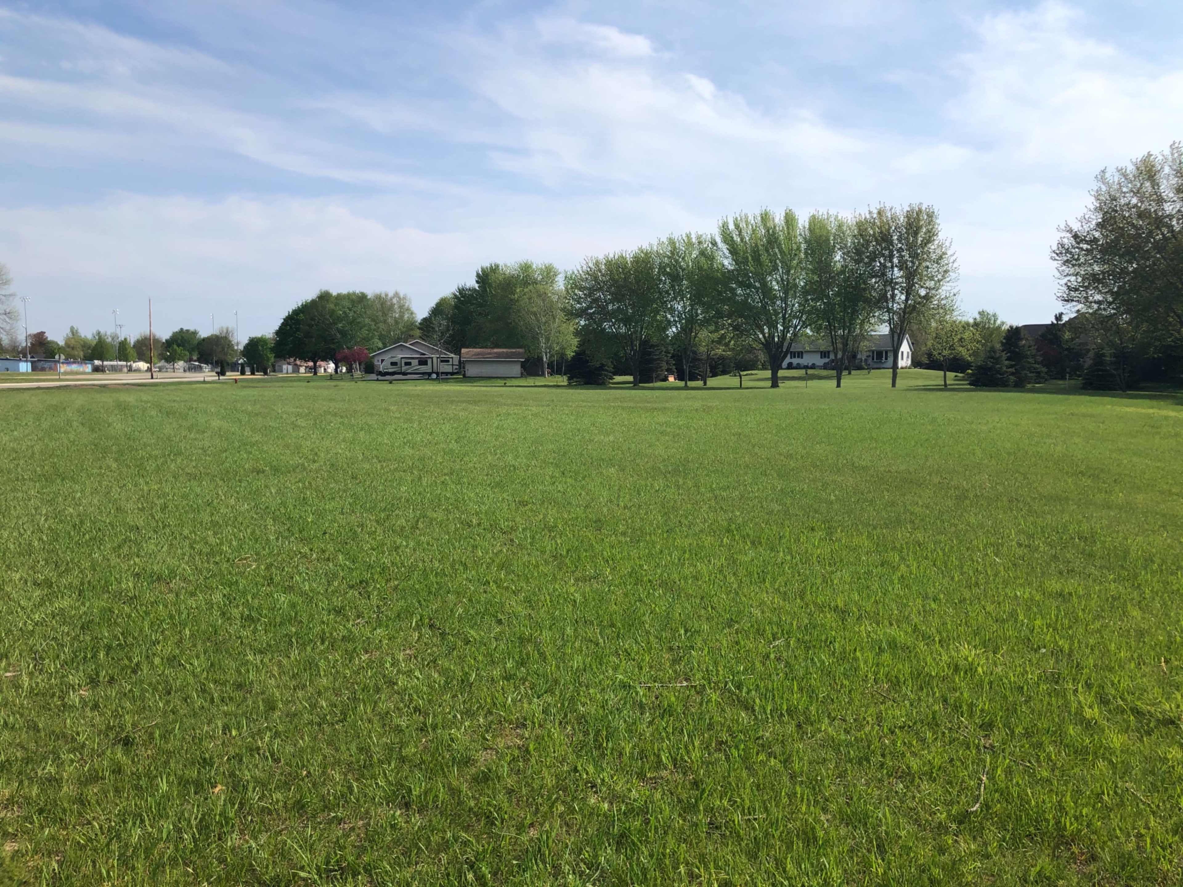 A vast green field with a few trees and houses lining the background under a partly cloudy sky.