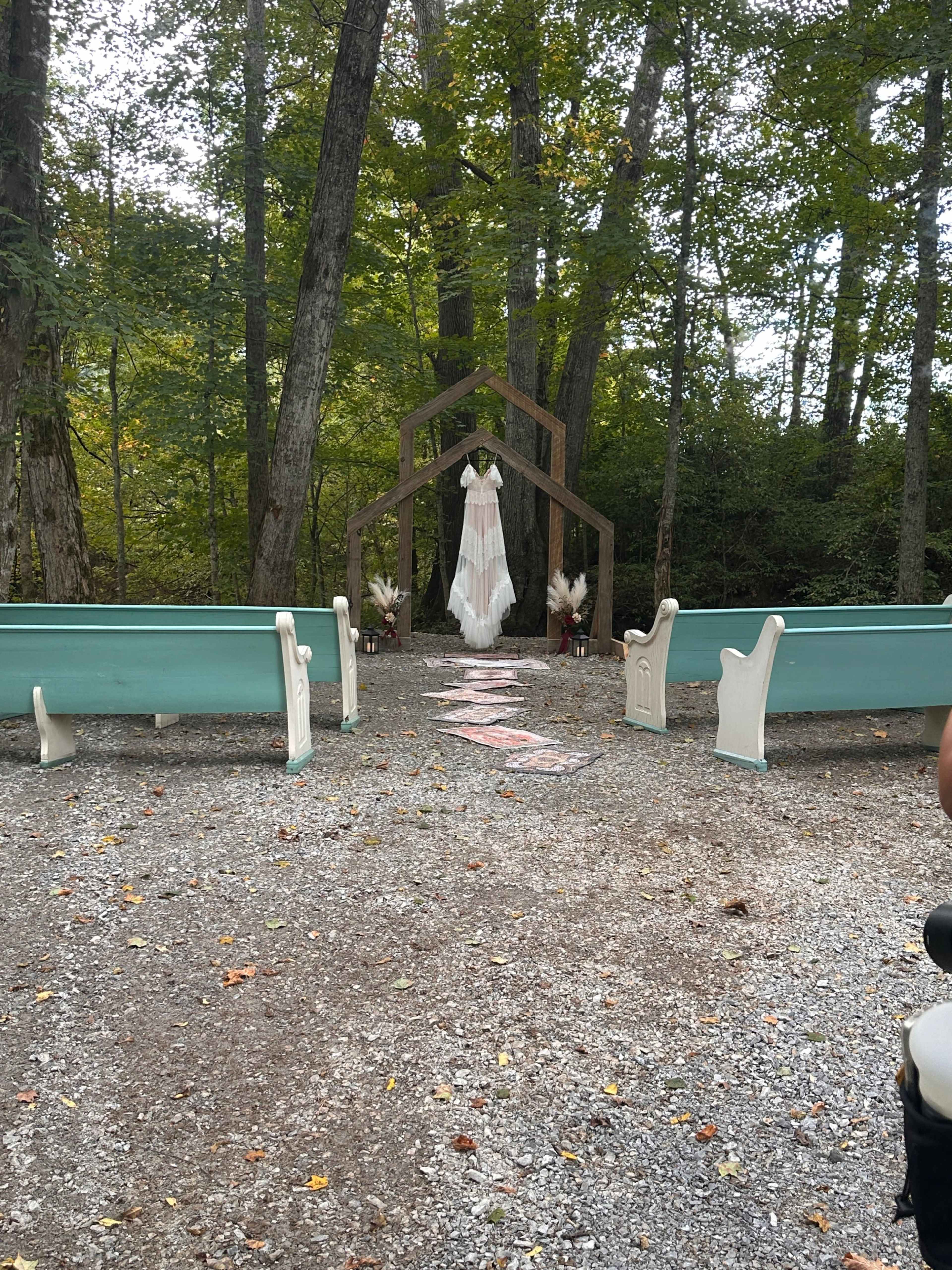 A rustic outdoor wedding setup features a wooden arch adorned with white fabric, flanked by benches on a gravel path surrounded by trees.