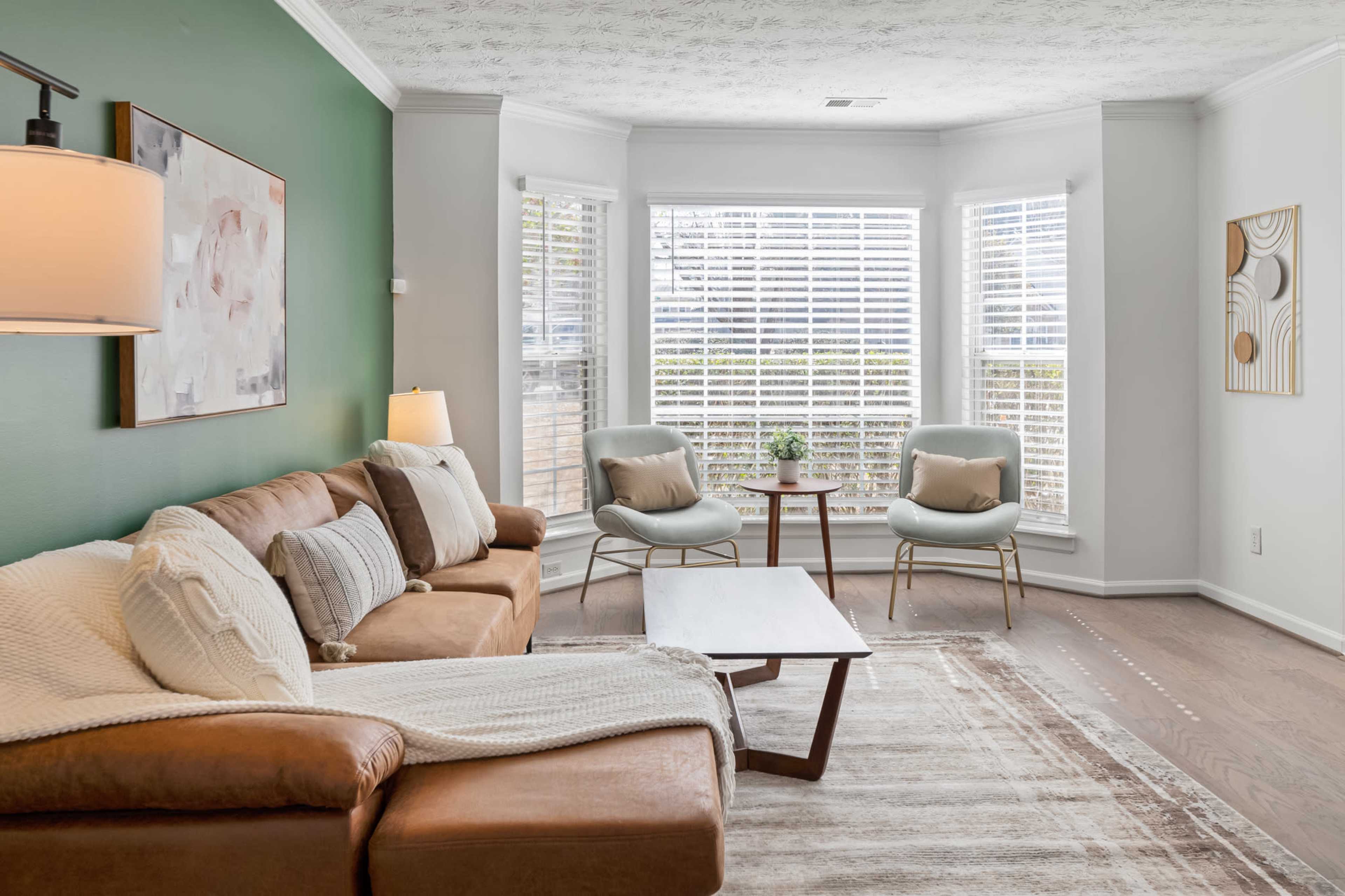 A cozy living room features a brown leather sofa, light blue armchairs, a wooden coffee table, and large windows with white blinds.