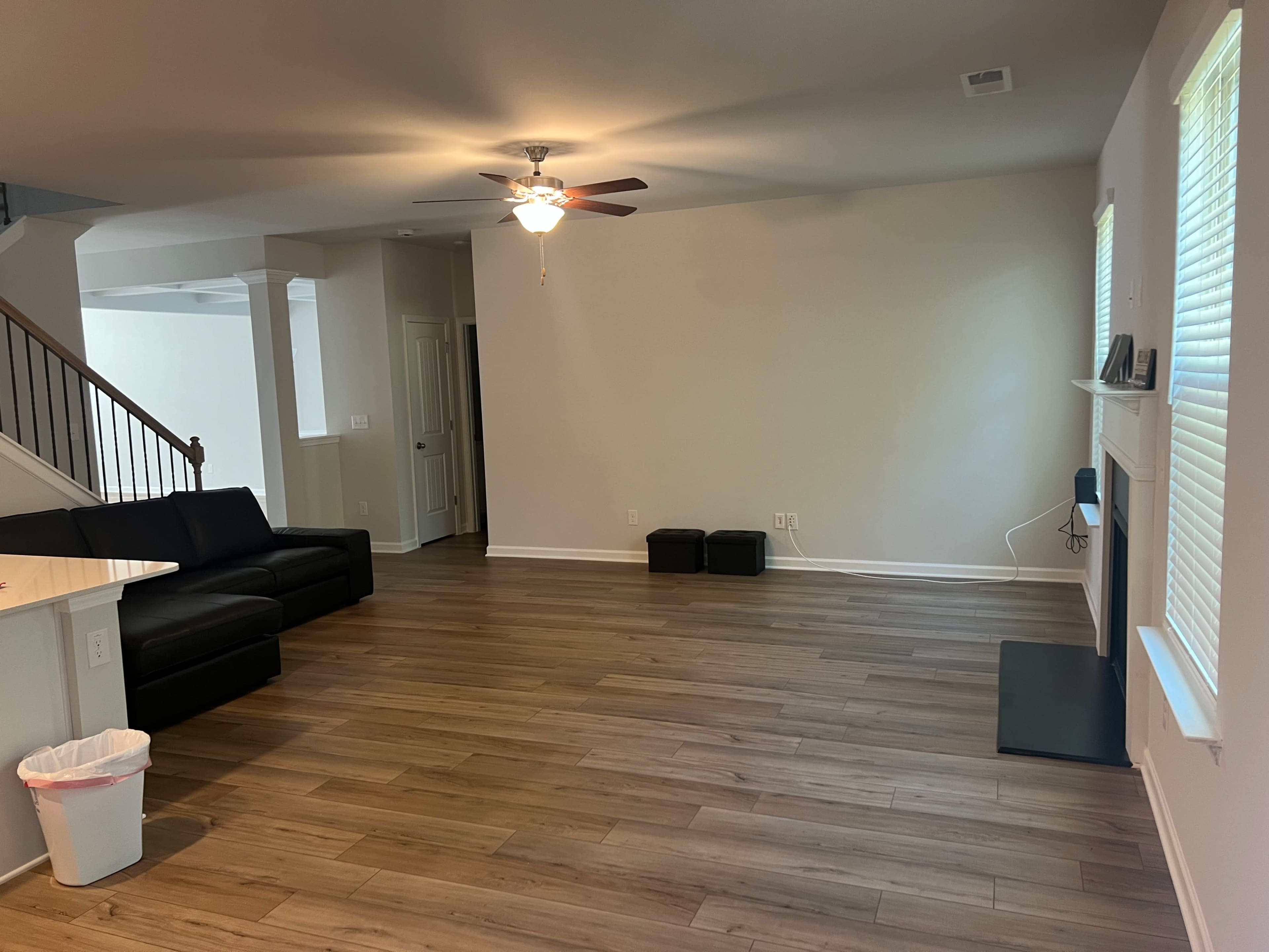 A spacious living room with light wood flooring, a black sofa, a ceiling fan, and a few windows allowing natural light.