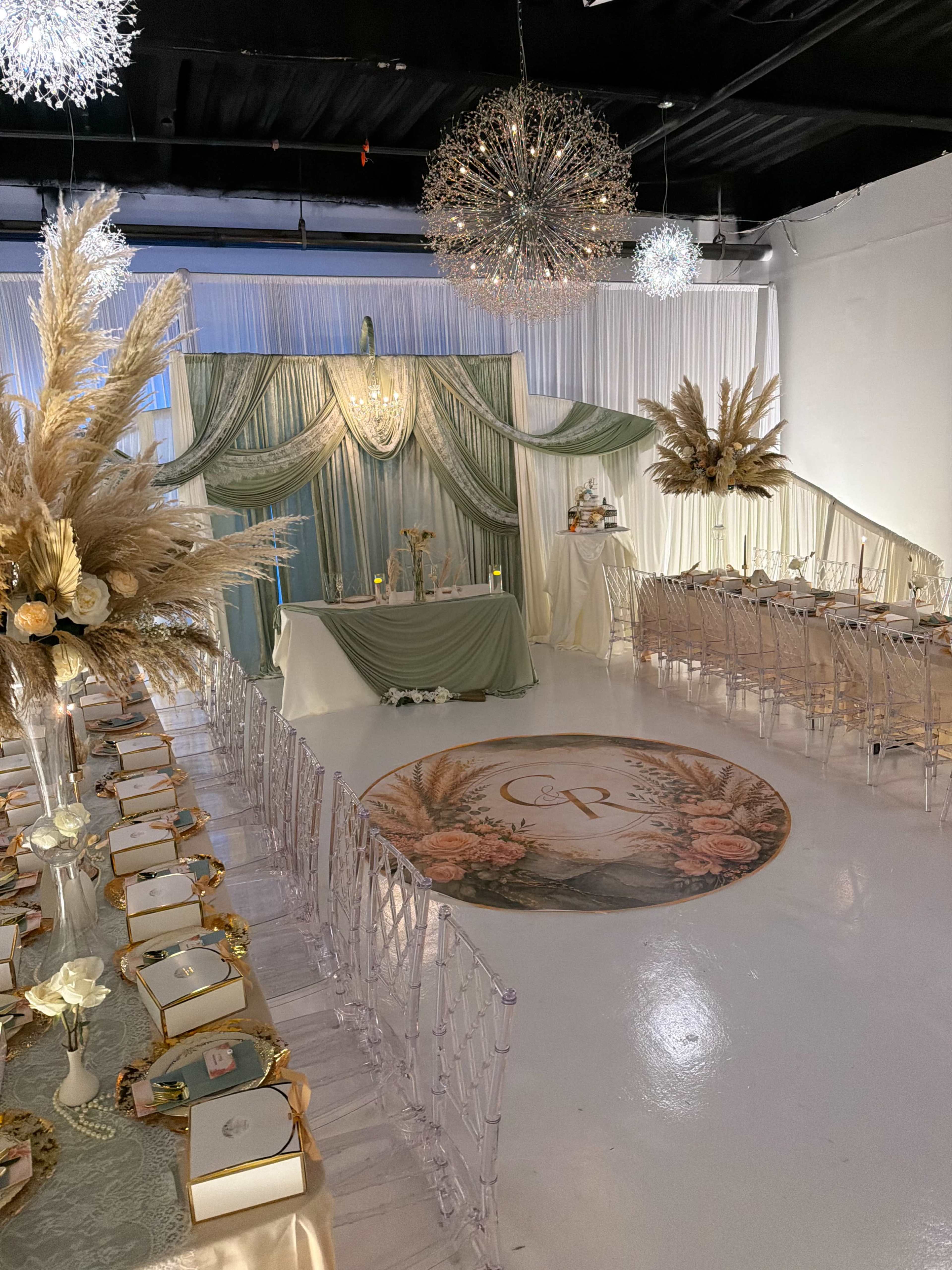 The image shows an elegantly decorated event space with a long banquet table, adorned with gold accents and floral centerpieces, against a backdrop of draped fabric and stylish chandeliers.