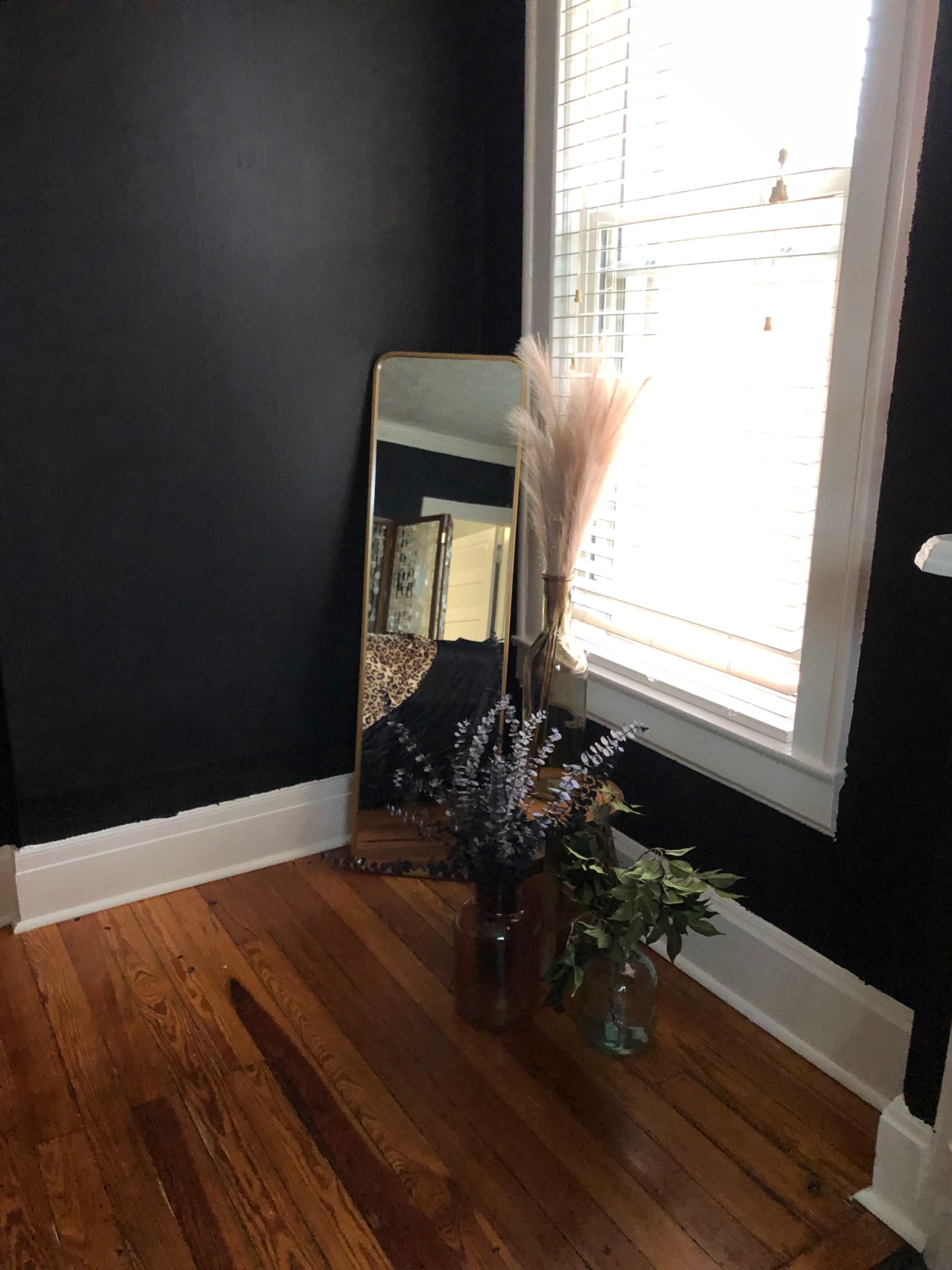 A tall mirror stands beside a window adorned with blinds, reflecting a corner filled with decorative plants in glass vases on a wooden floor.