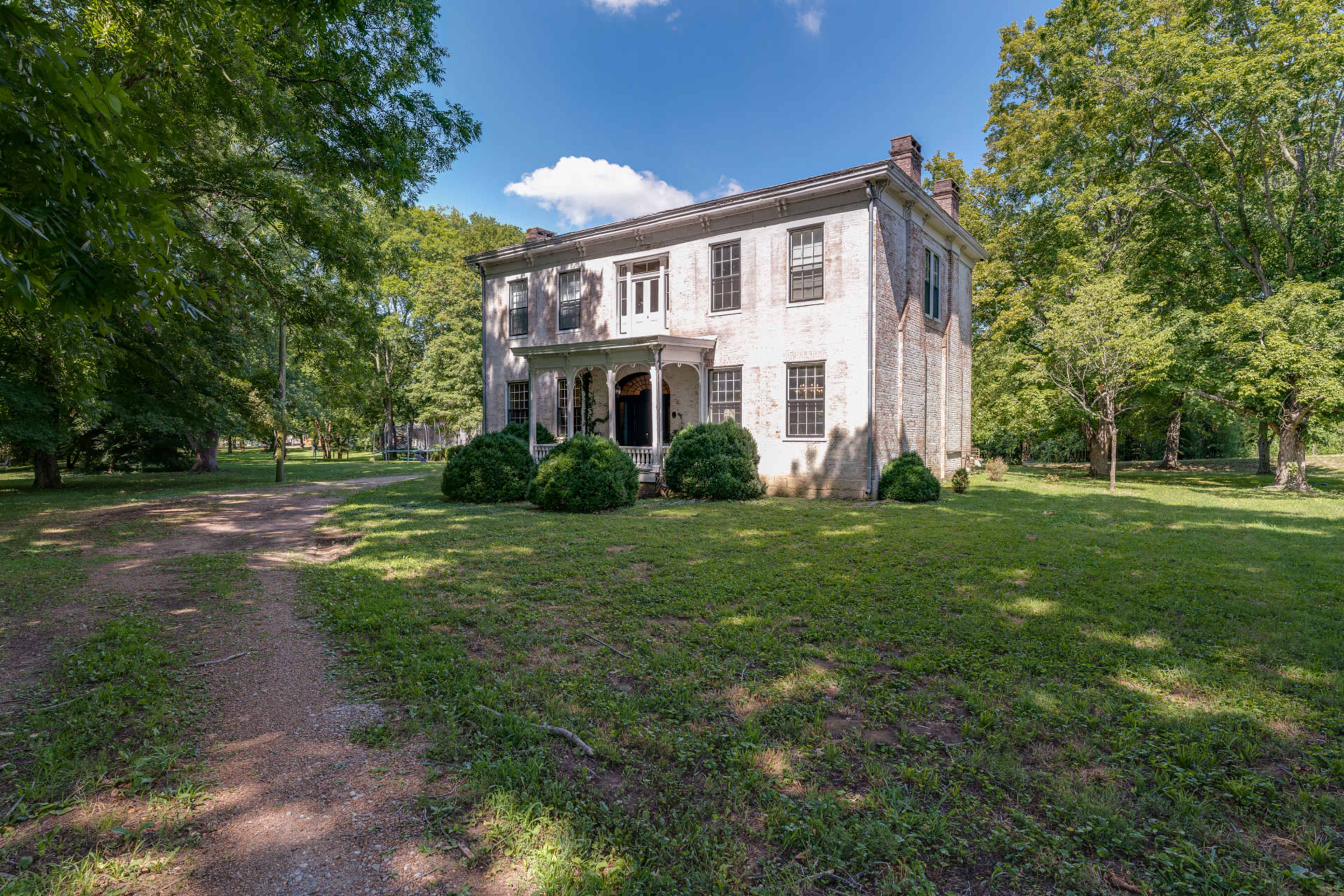 A two-story, weathered brick house is surrounded by greenery and stands on a gravel path.