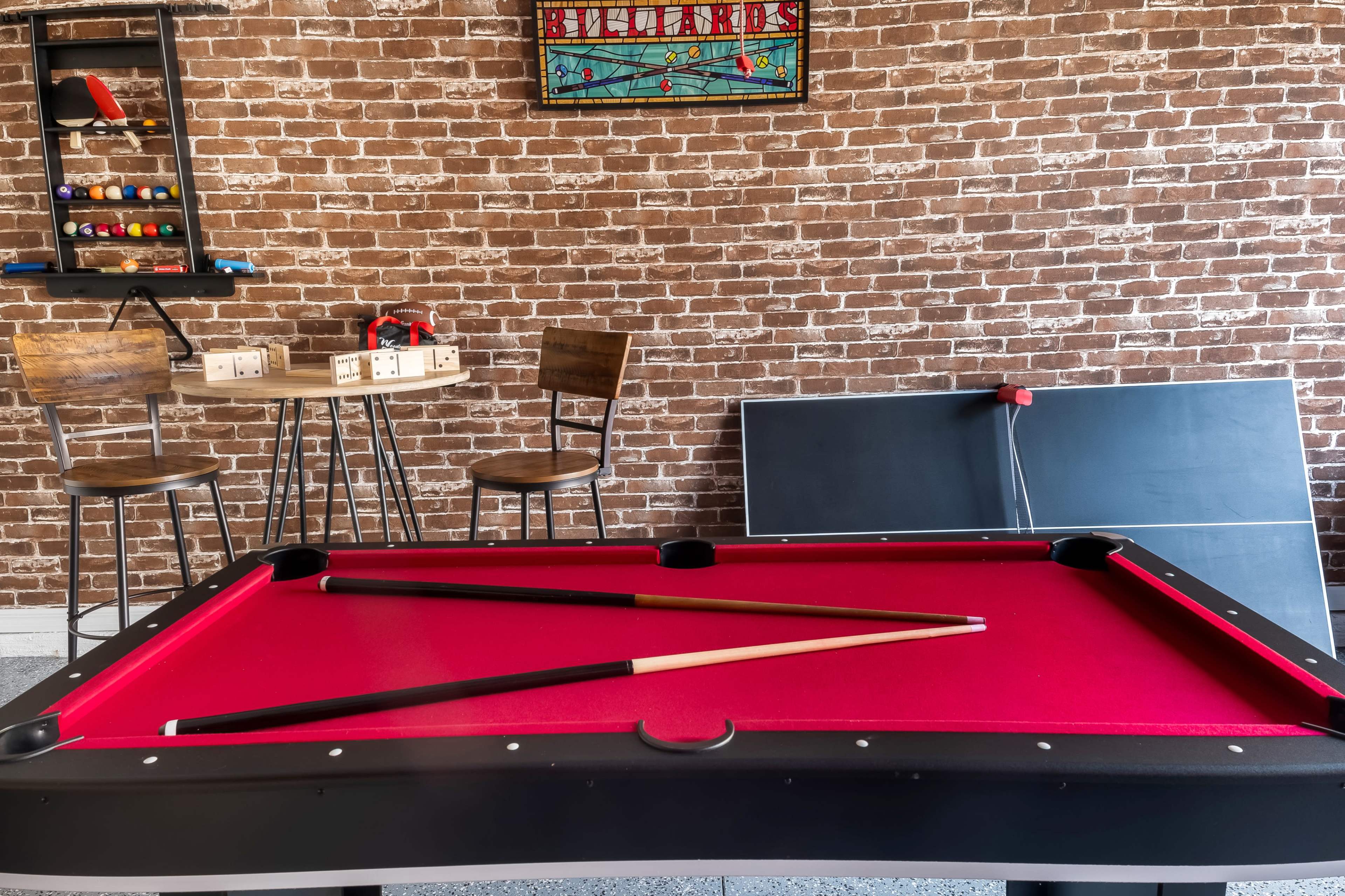 A red-felt billiard table is centered in a room with a brick wall, featuring a small table and chairs in the background.